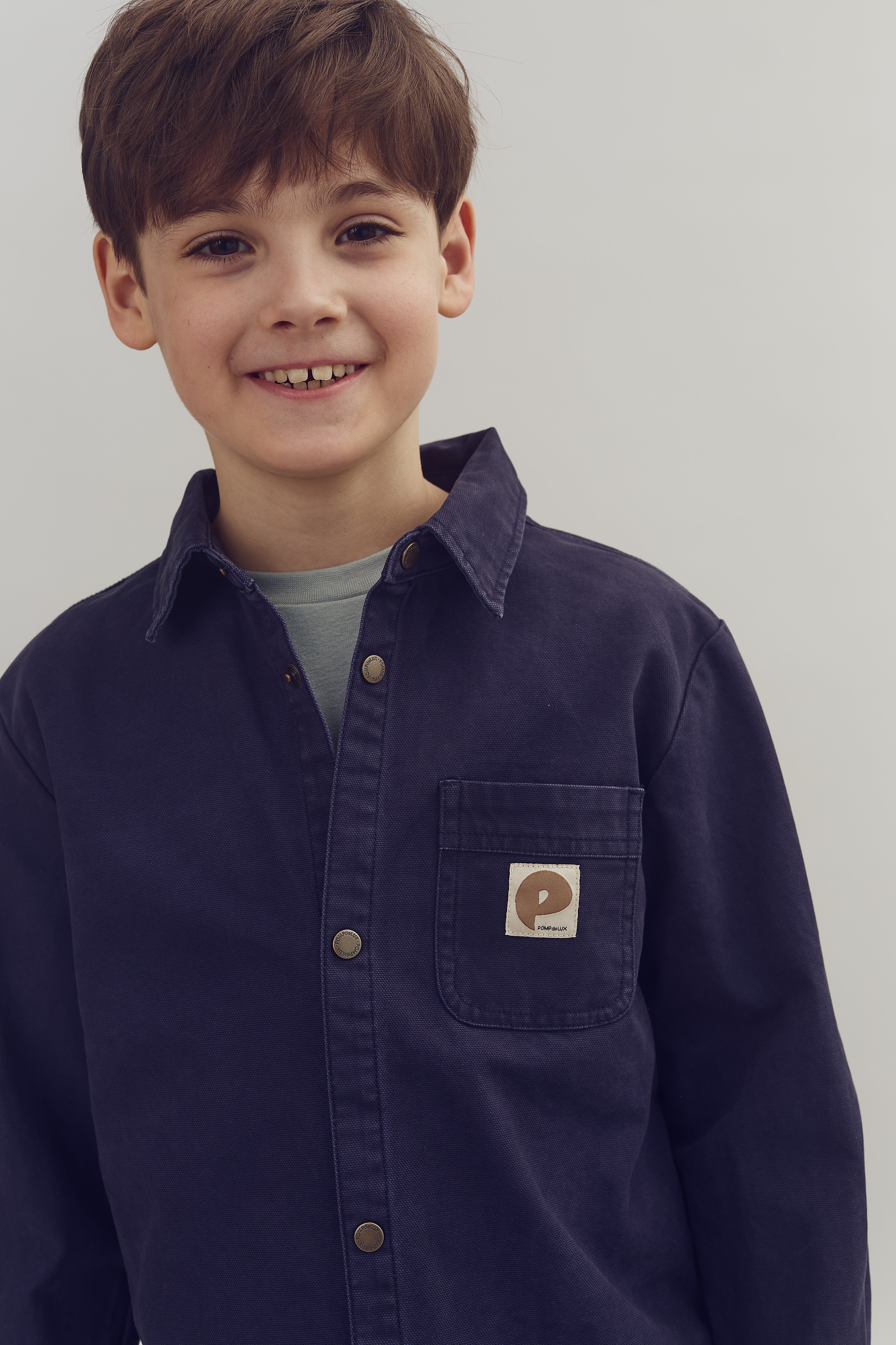A boy smiles at the camera, wearing a dark blue button-up shirt with a logo "P" on the chest pocket, against a plain light background.