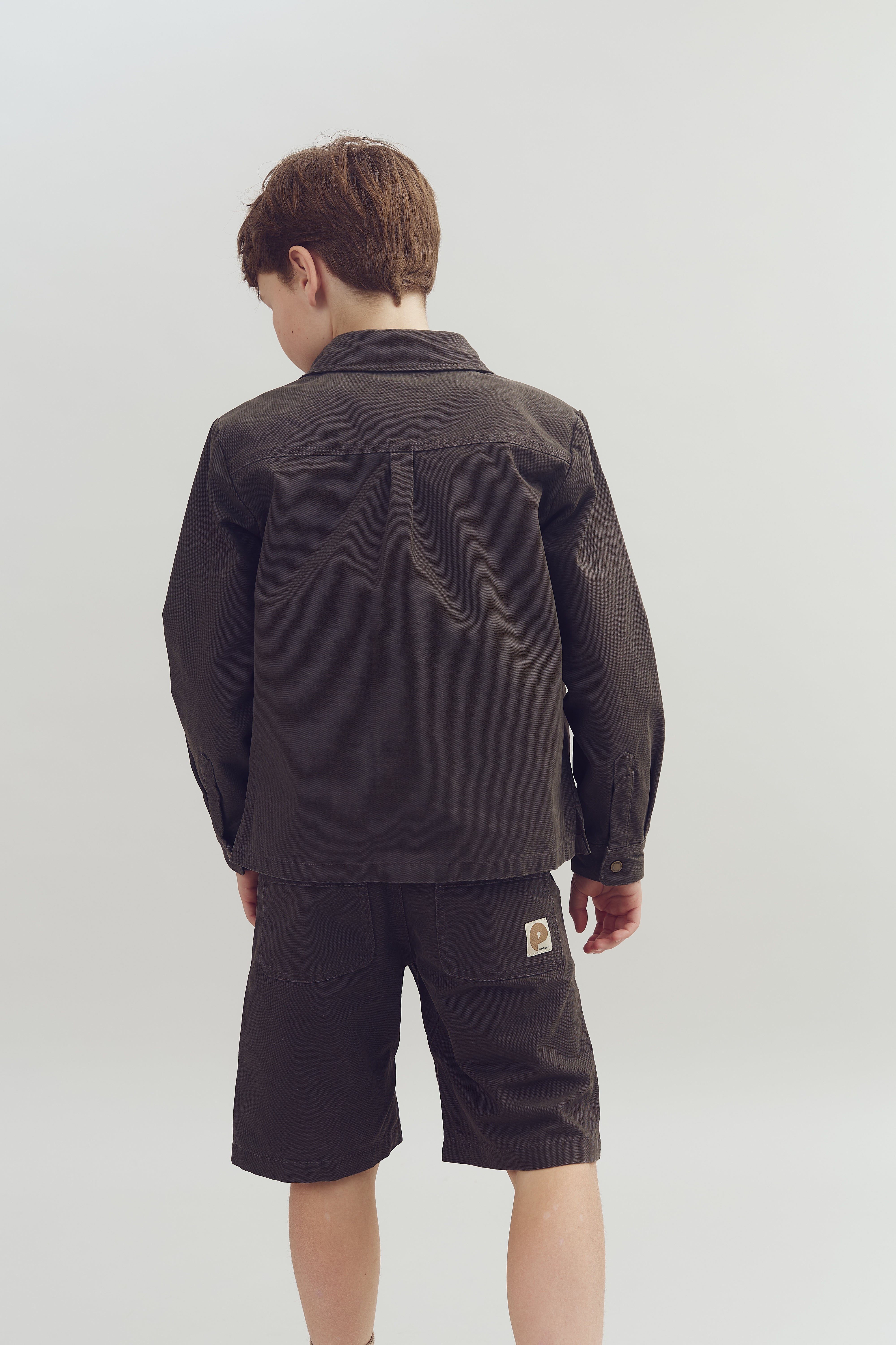 A child stands facing away, wearing a dark jacket and shorts with a logo on the pocket. The background is plain and light-colored, emphasizing the clothing.