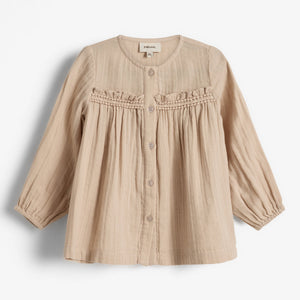 Beige Kinderbluse mit Knöpfen, Volants und langen Ärmeln; auf einem weißen Hintergrund präsentiert. Das Etikett im Nacken zeigt „Poudre Organic“.