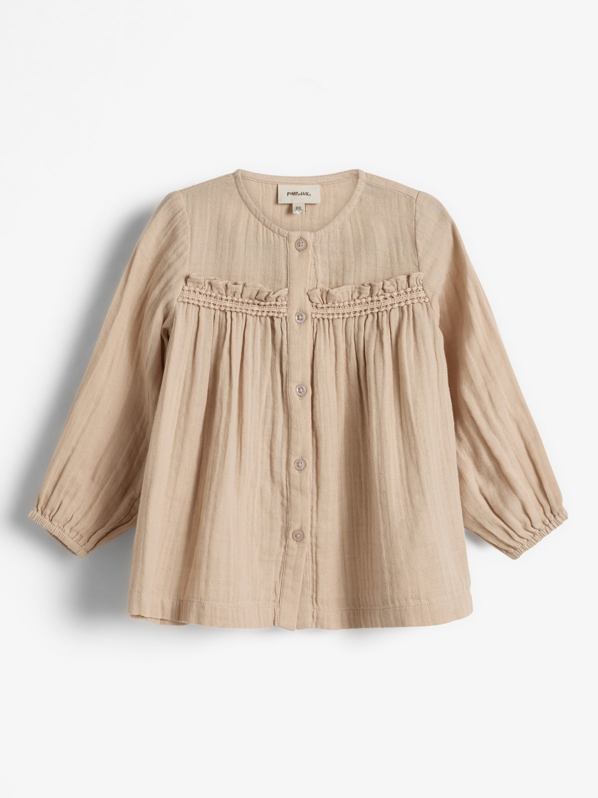 Beige Kinderbluse mit Knöpfen, Volants und langen Ärmeln; auf einem weißen Hintergrund präsentiert. Das Etikett im Nacken zeigt „Poudre Organic“.