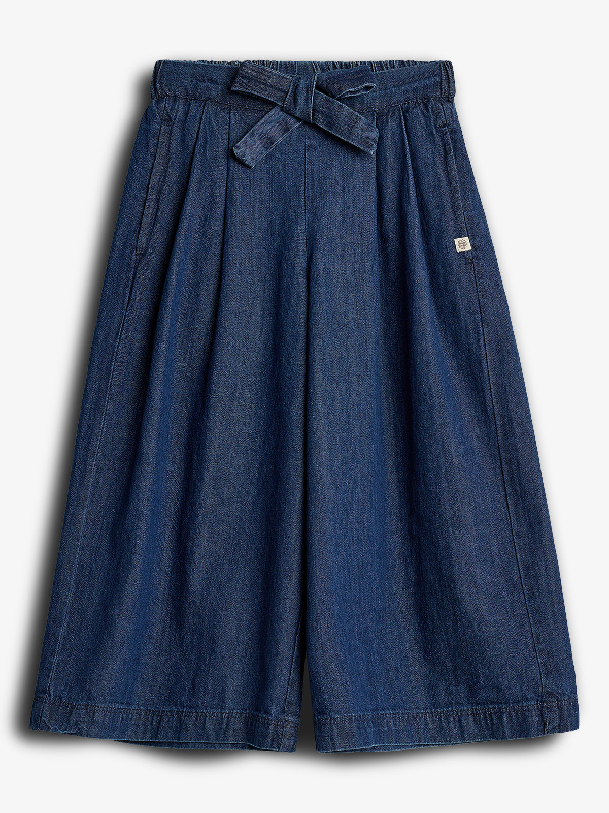 Wide-legged denim culottes feature a tied waist and deep pleats, displayed against a plain white background. A small logo tag is attached on the right side.