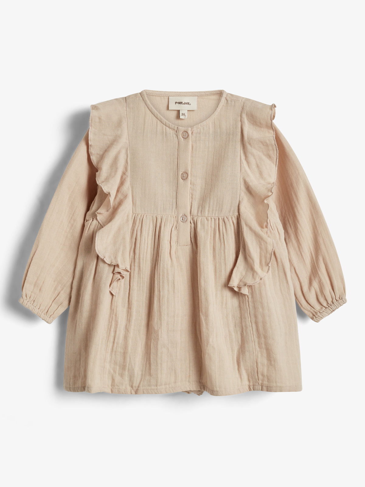 Eine beige Kinderbluse mit langen Ärmeln und Rüschen an den Schultern. Sie hat vorne drei sichtbare Knöpfe. Im neutralen Hintergrund hängt ein Etikett mit „POMPdeLUX“ und „86“.