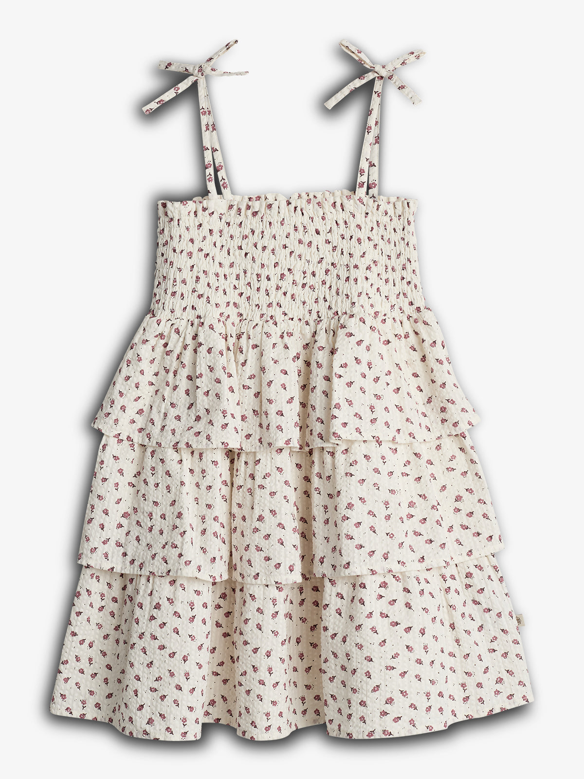 Sleeveless dress with tied shoulder straps, features a smocked bodice and tiered ruffles. Covered in small red floral patterns on a white background, displayed against a plain backdrop.