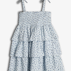 A light blue, sleeveless dress with a smocked bodice and tiered ruffle skirt, adorned with small blue floral patterns, has tied shoulder straps. Displayed against a plain white background.