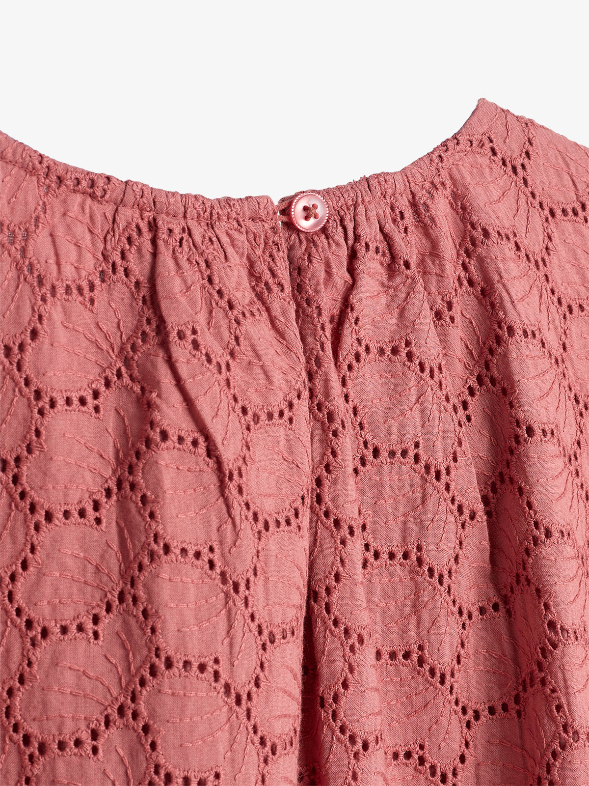 Pink top buttoned at the back, featuring intricate eyelet embroidery in a circular leaf pattern. The fabric is gathered near the neckline, creating a delicate texture against a plain background.