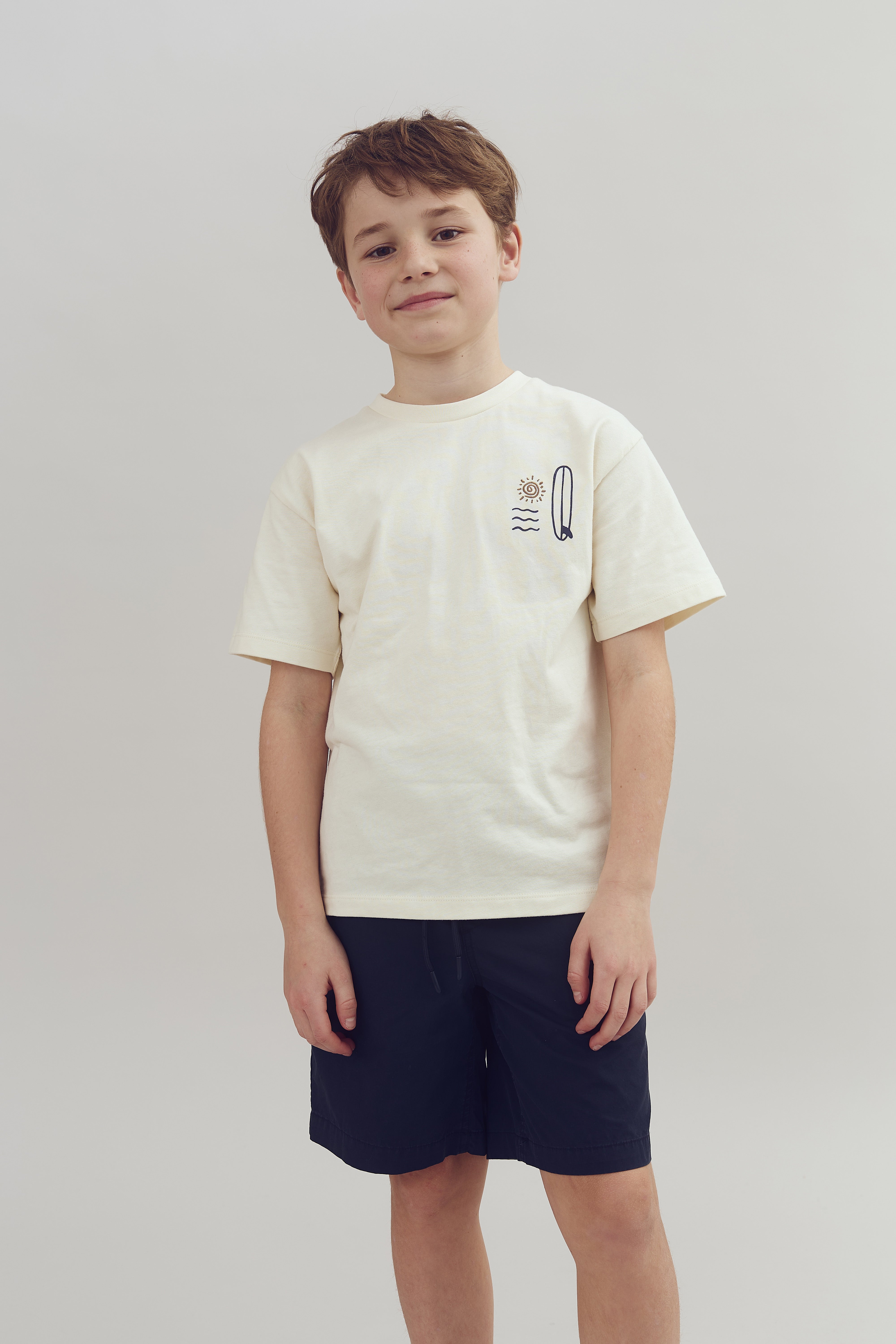 A boy stands smiling slightly, wearing a cream-colored t-shirt with a sun, wave, and feather design, paired with navy shorts. The background is a plain, light gray color.