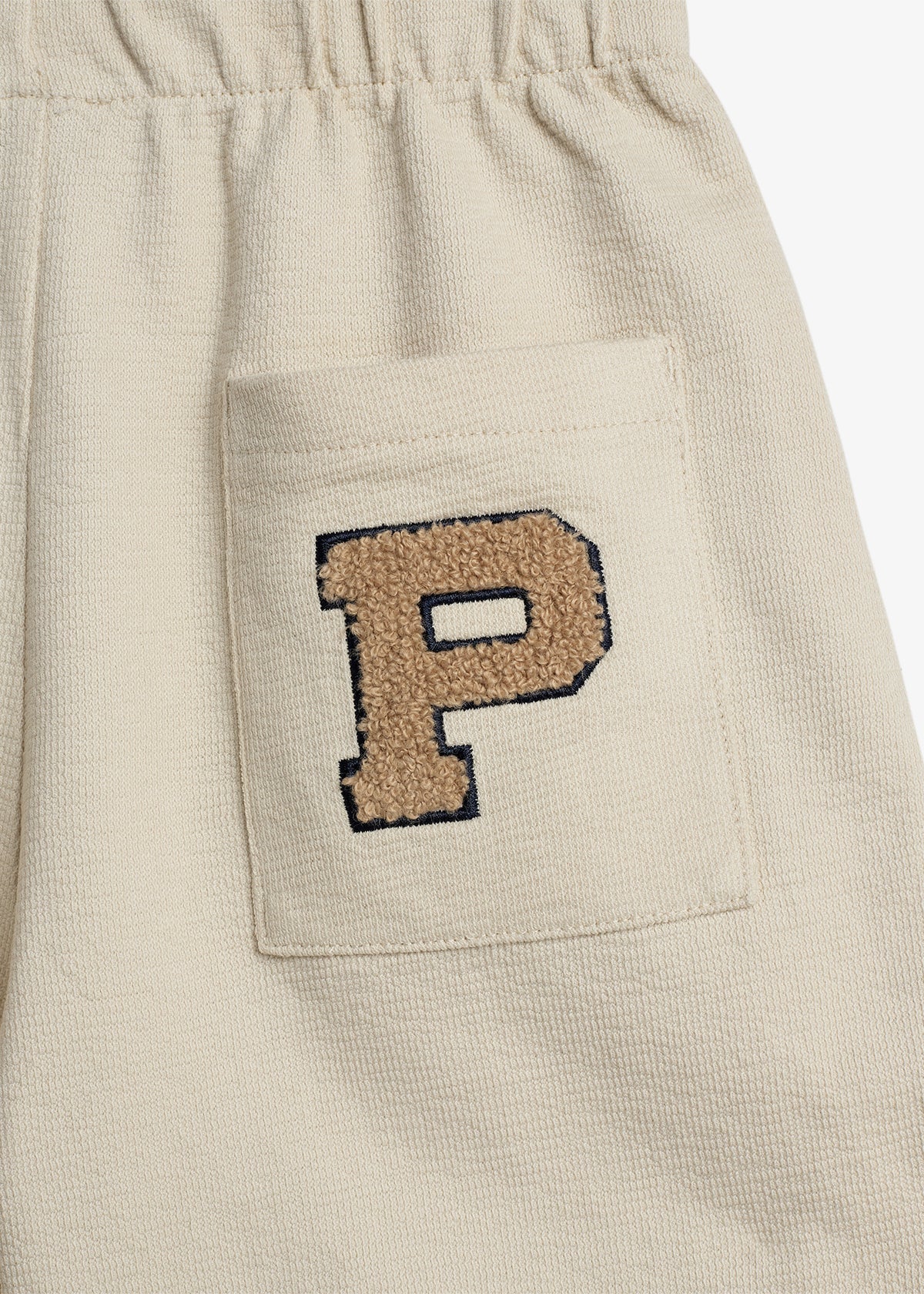 Beige shorts feature an embroidered patch with a large textured letter "P" in brown and black, located on a pocket. The fabric appears textured, and the shorts have an elastic waistband.