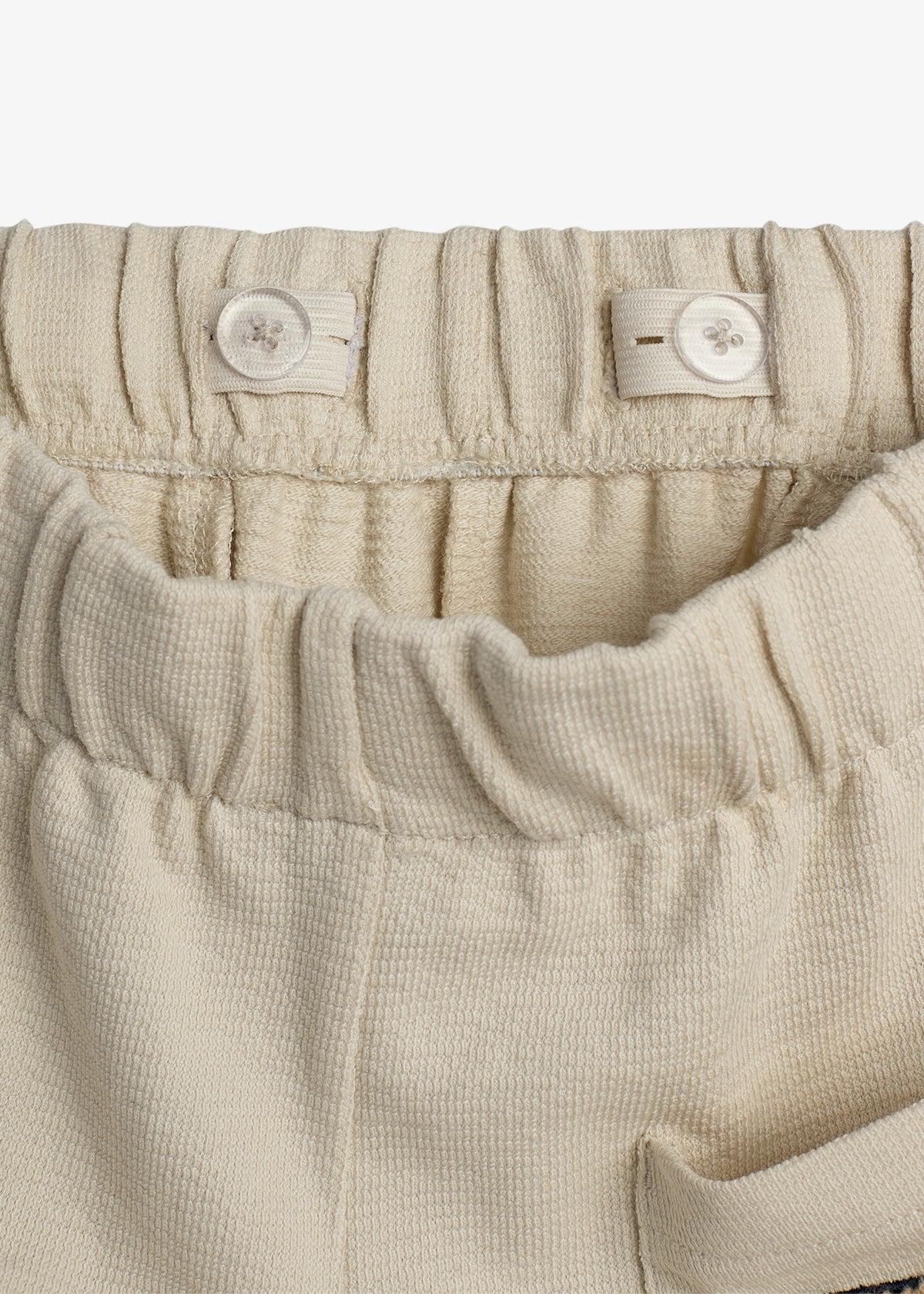 Cream-colored fabric waistband with elastic and adjustable button tabs, creating small gathers; features textured material, suggesting children's pants in a simple, neutral setting.