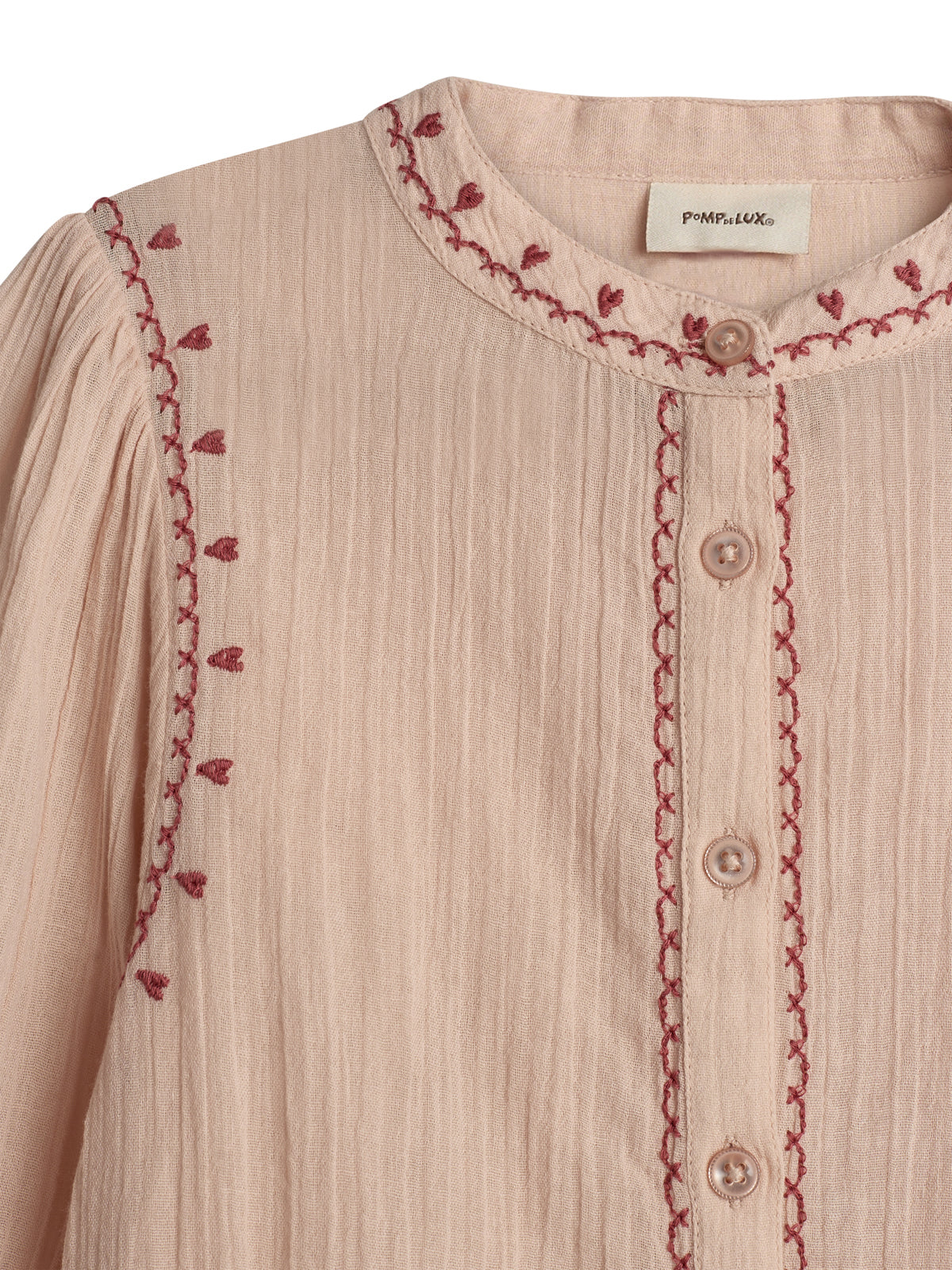 A light pink blouse with intricate red embroidery along the collar and sleeve edges, featuring a button-down design. The label reads "POMPdeLUX," and the fabric appears textured.