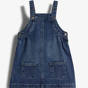 Jeanskleid mit Trägern, verziert mit Knöpfen und zwei aufgesetzten Taschen vorne. Liegt auf einem weißen Hintergrund.