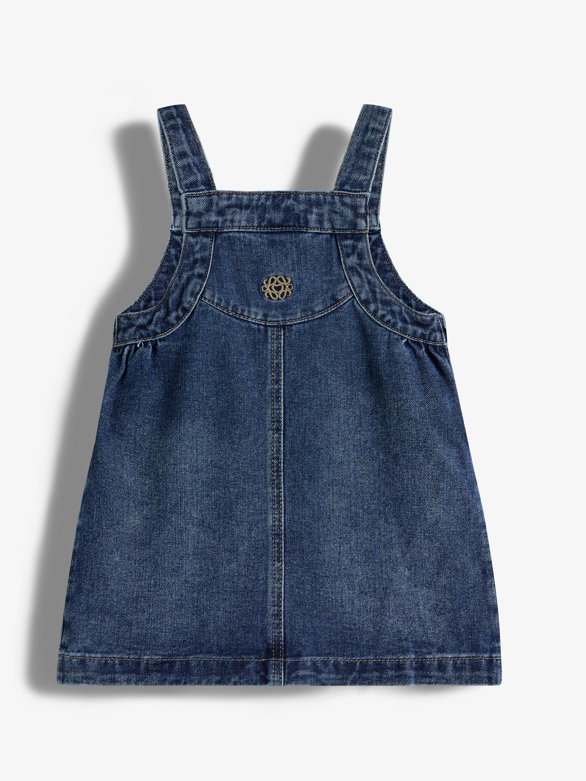 Ein Jeanskleid mit breiten Trägern liegt flach auf einem weißen Hintergrund. Es hat eine dekorative, goldene Plakette auf der Vorderseite in der Mitte.