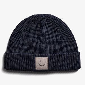 A dark blue knitted beanie displays a small, beige patch with a smiley face and the text "POMPdeLUX" on its folded brim, set against a plain white background.