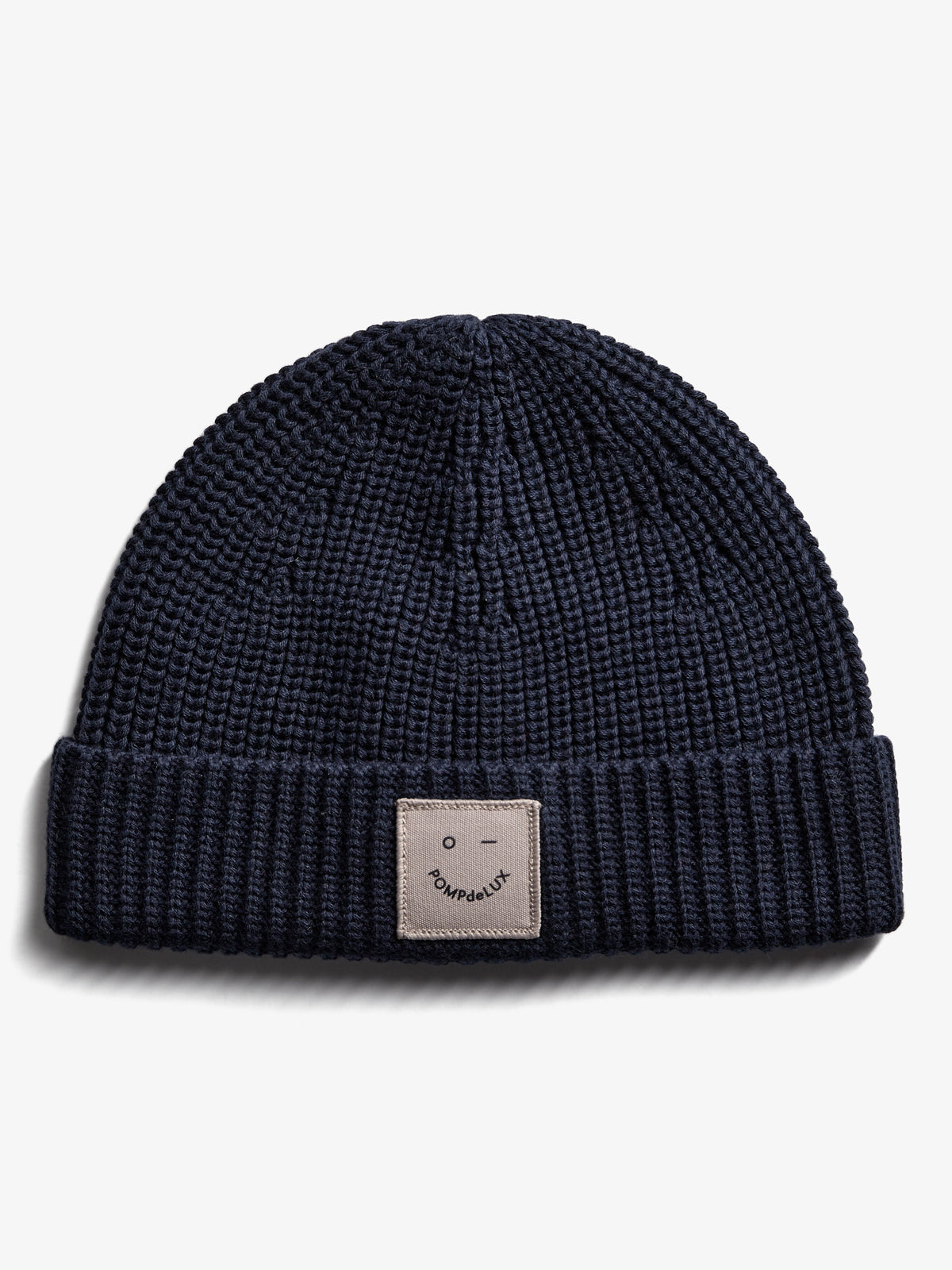 A dark blue knitted beanie displays a small, beige patch with a smiley face and the text "POMPdeLUX" on its folded brim, set against a plain white background.