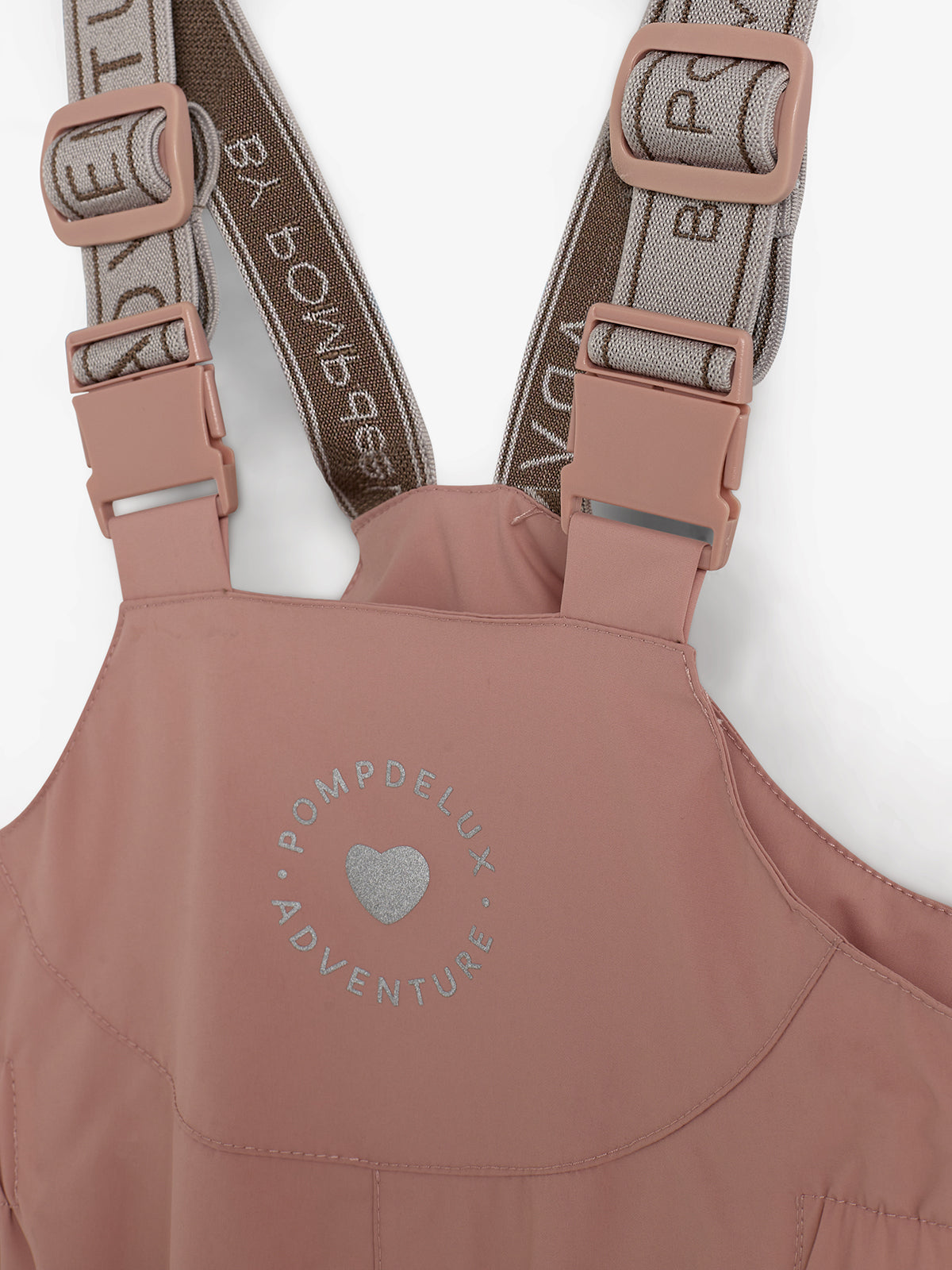 Rosa Trägerhose mit Kunststoffschnallen aus Kunststoff, verstellbaren elastischen Riemen und der Aufschrift „POMPDELUX ADVENTURE“ mit einem silbernen Herzsymbol vorne, vor einem weißen Hintergrund.
