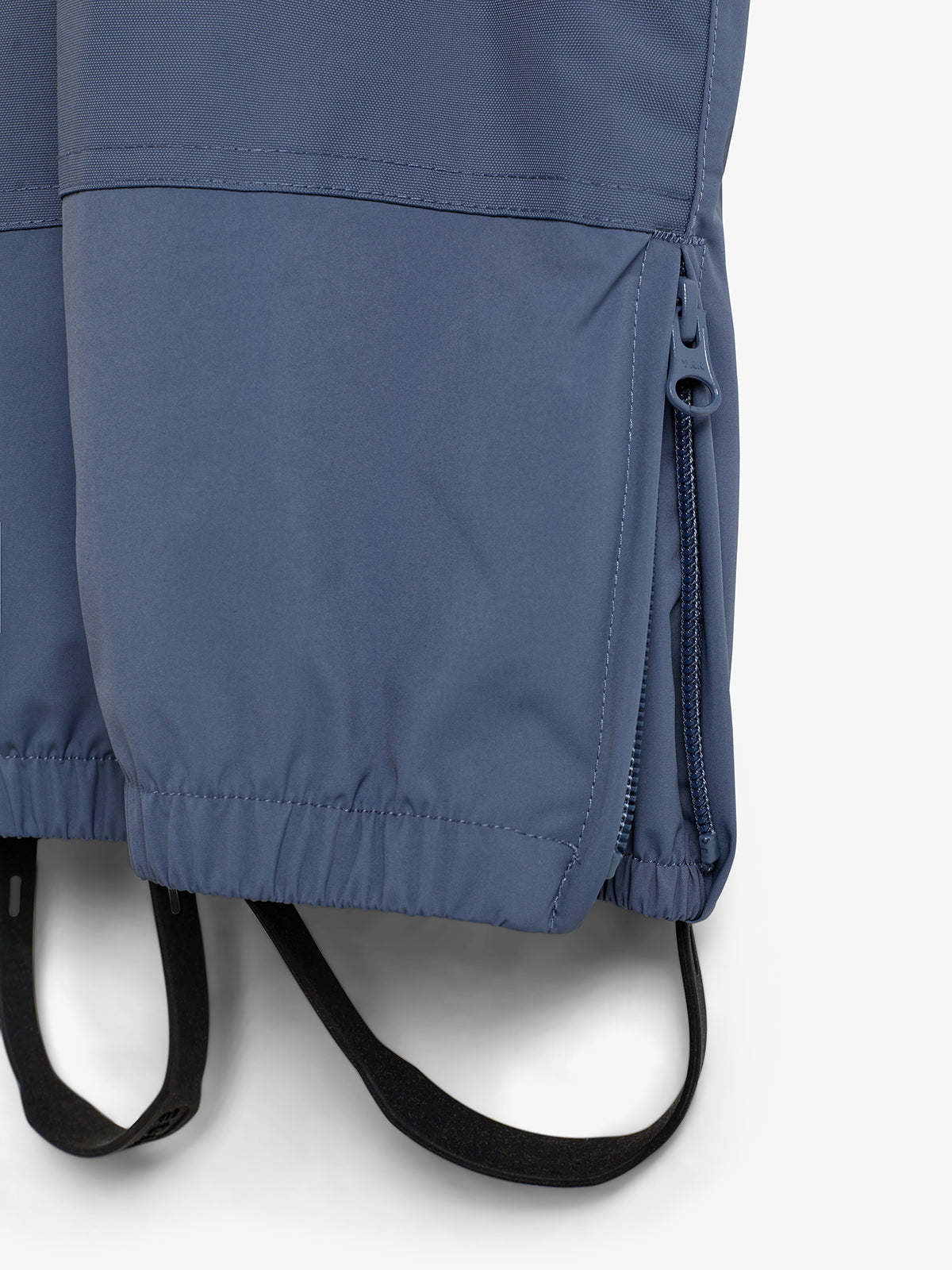 Eine blaue Hose mit Reißverschluss seitlich und Gummizug unten hängt, umgeben von einem weißen Hintergrund. Elastische Fußschlaufen sind sichtbar.
