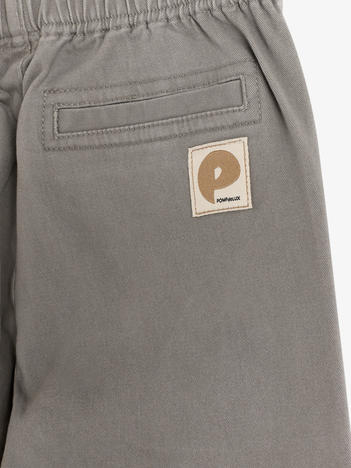 Eine graue Hose mit aufgenähter Tasche zeigt ein Etikett mit dem Buchstaben "P" und dem Text "POMPdeLUX", umgeben von einem hellen Hintergrund.