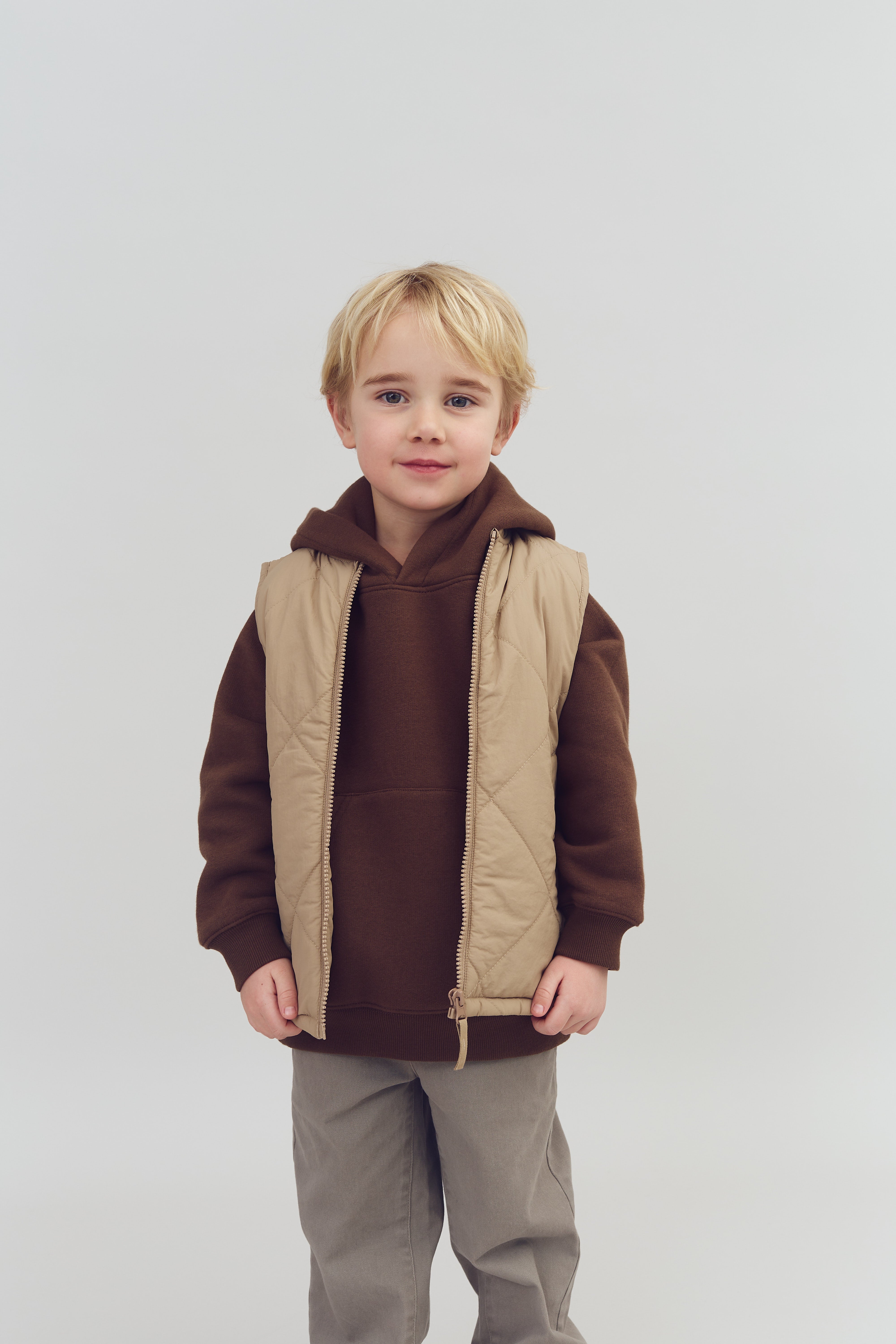 A young child stands smiling, wearing a brown hoodie and beige vest, with hands by their side. The background is a simple, light gray, providing a neutral setting.