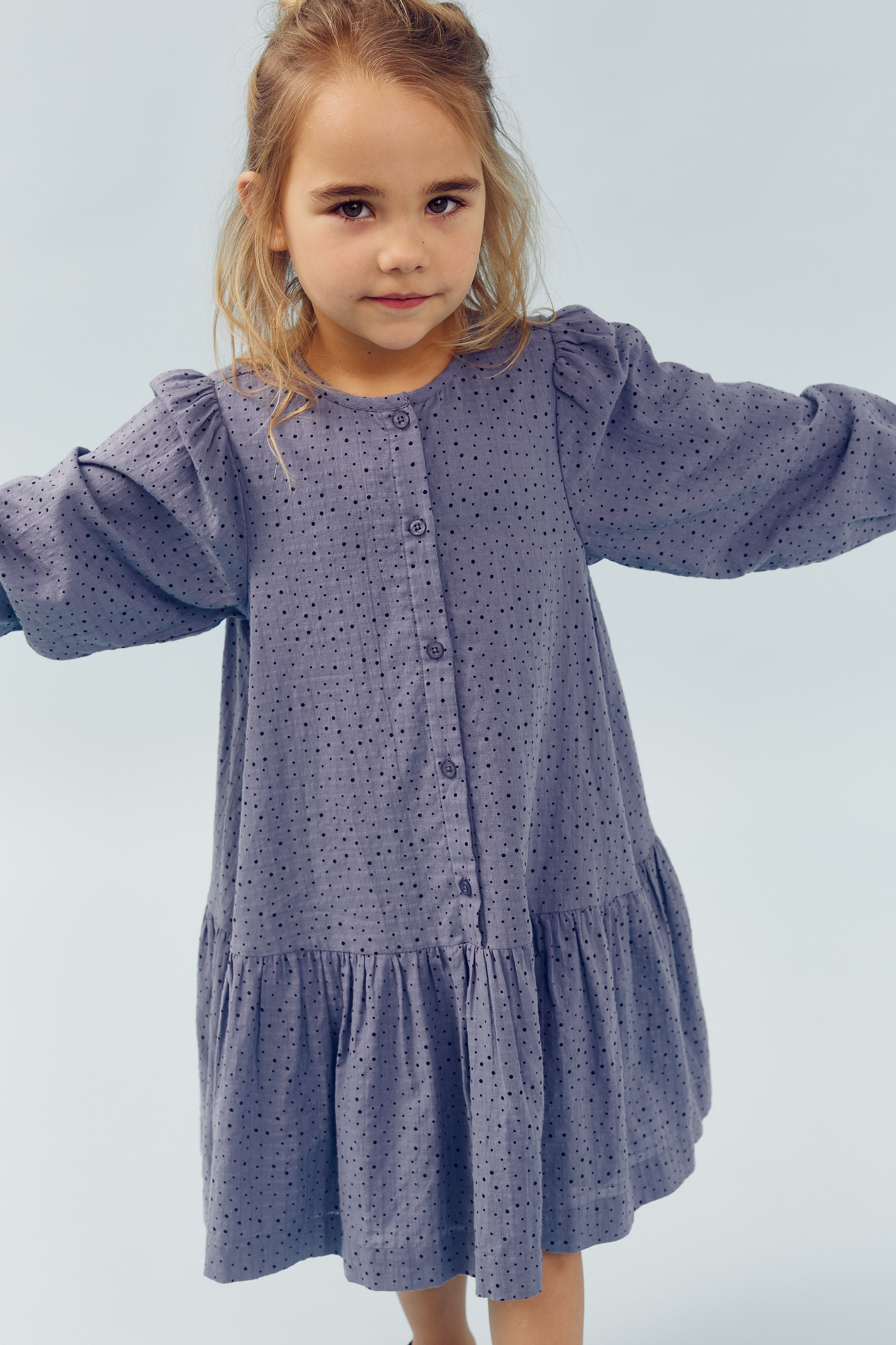 A young child stands in a long-sleeve, button-up, purple dress with a polka dot pattern, arms outstretched, against a light blue background.