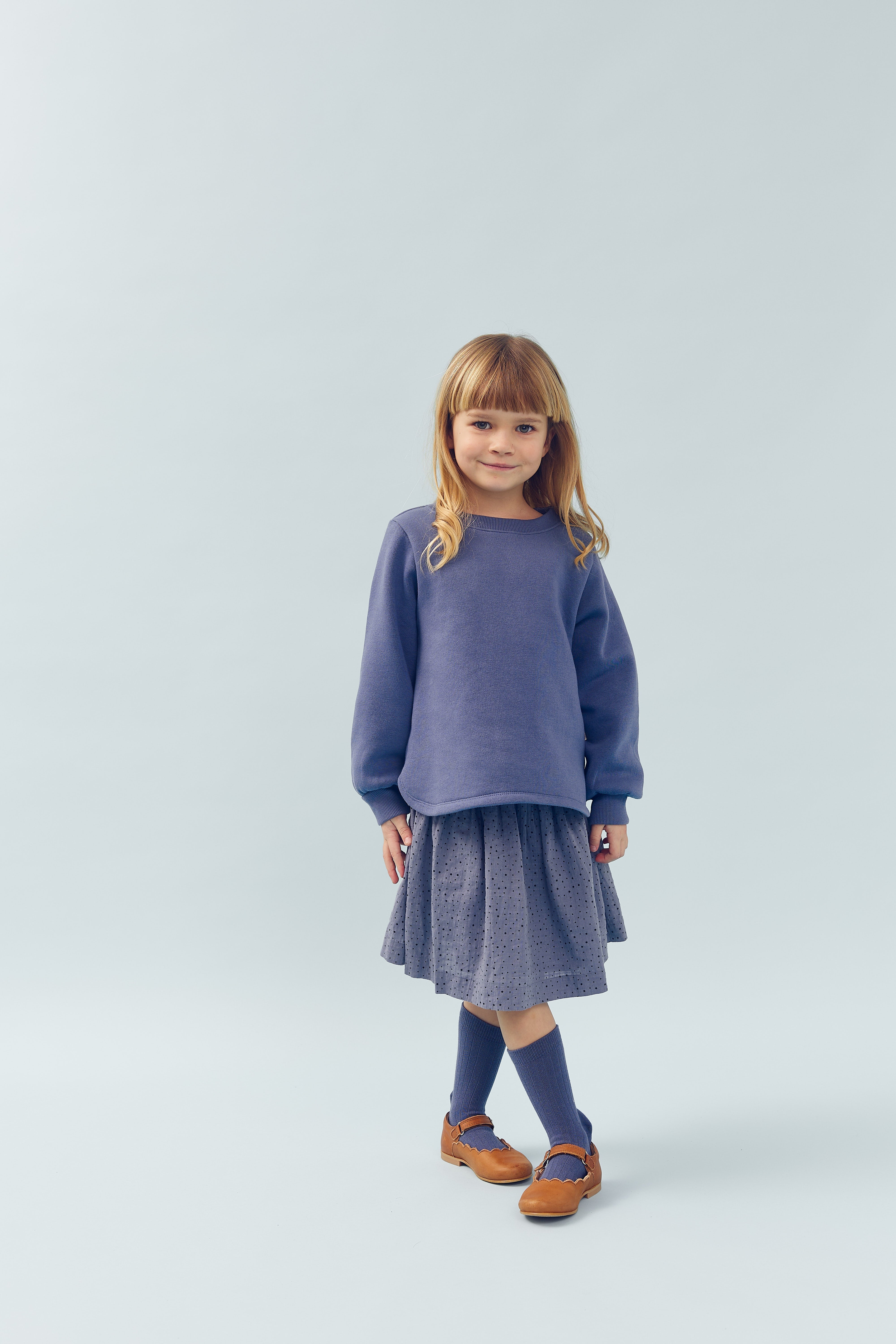 Ein Kind steht lächelnd; es trägt einen blauen Pullover, einen grauen Rock, blaue Socken und braune Schuhe. Der Hintergrund ist schlicht und hell.