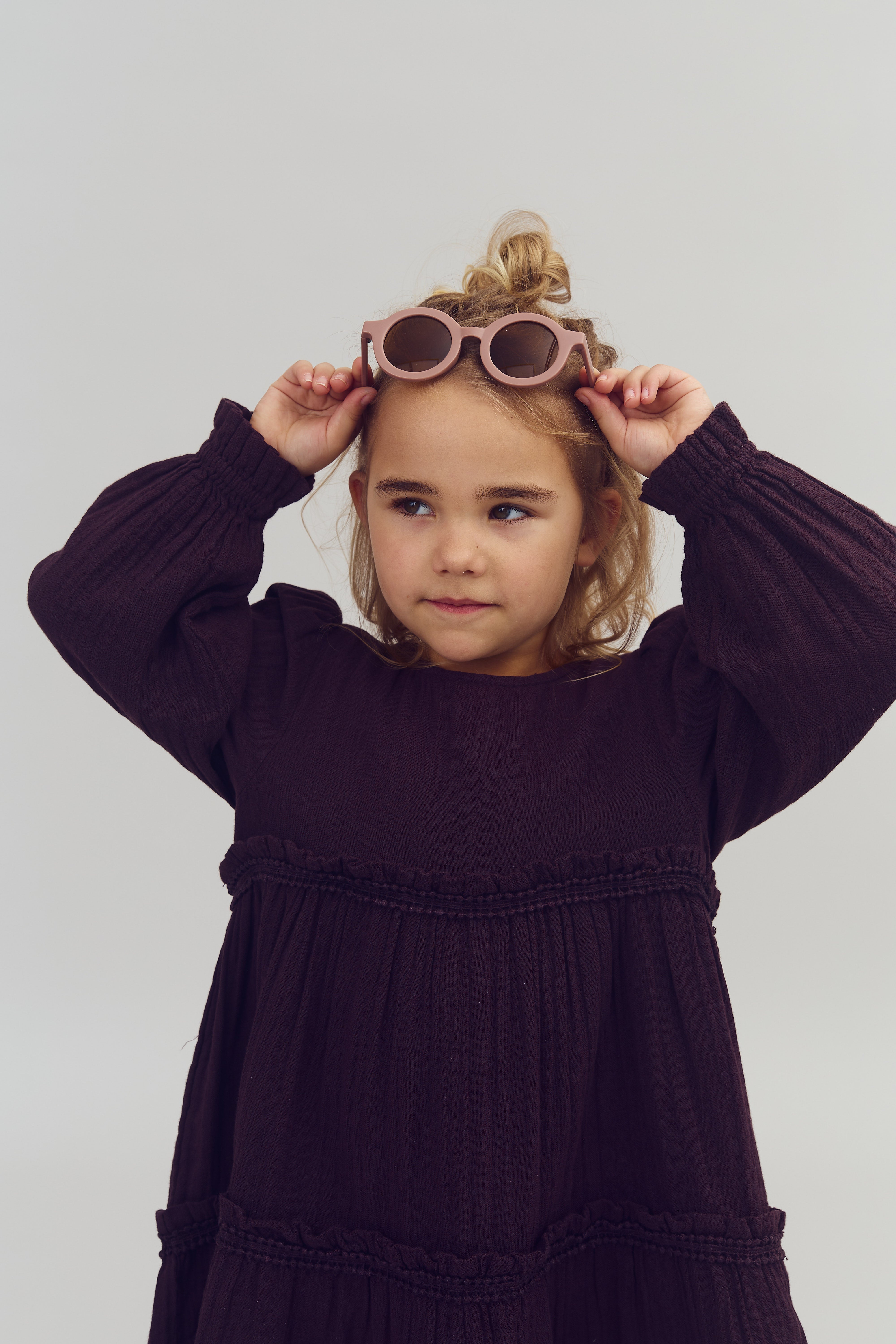 Ein Kind, trägt eine dunkellila Kleid, hebt eine rosa Sonnenbrille über den Kopf. Es sieht nach rechts, vor einem neutralen, grauen Hintergrund.