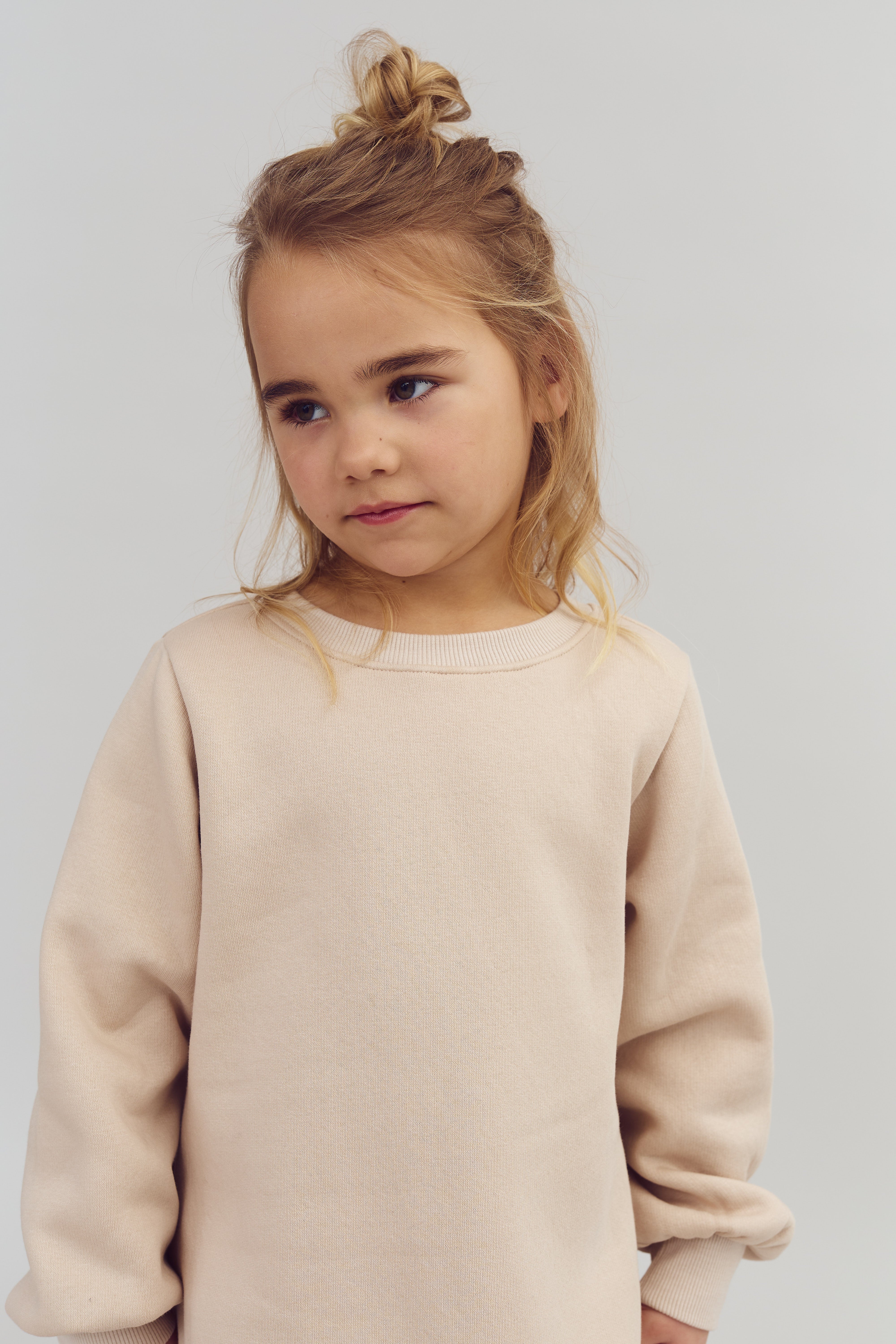 Ein Kind mit blonden Haaren im lockeren Dutt steht und schaut nachdenklich zur Seite. Es trägt einen beigefarbenen Pullover in einem neutralen, hellen Raum.