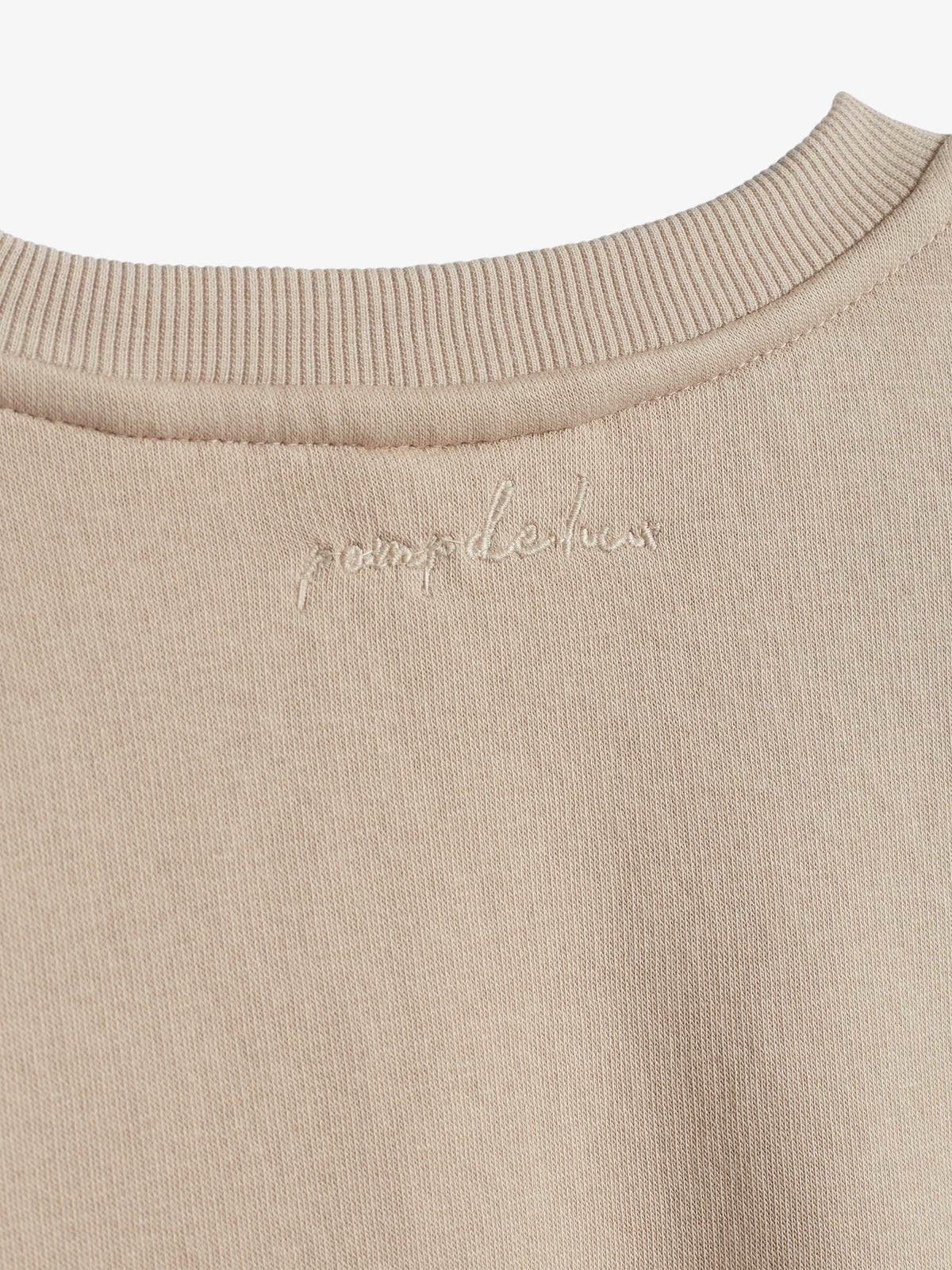 Beiger Pullover mit gesticktem Text "pomp de lux" am Kragen, präsentiert auf weißem Hintergrund. Der Kragen hat eine gerippte Struktur und der Stoff wirkt weich.