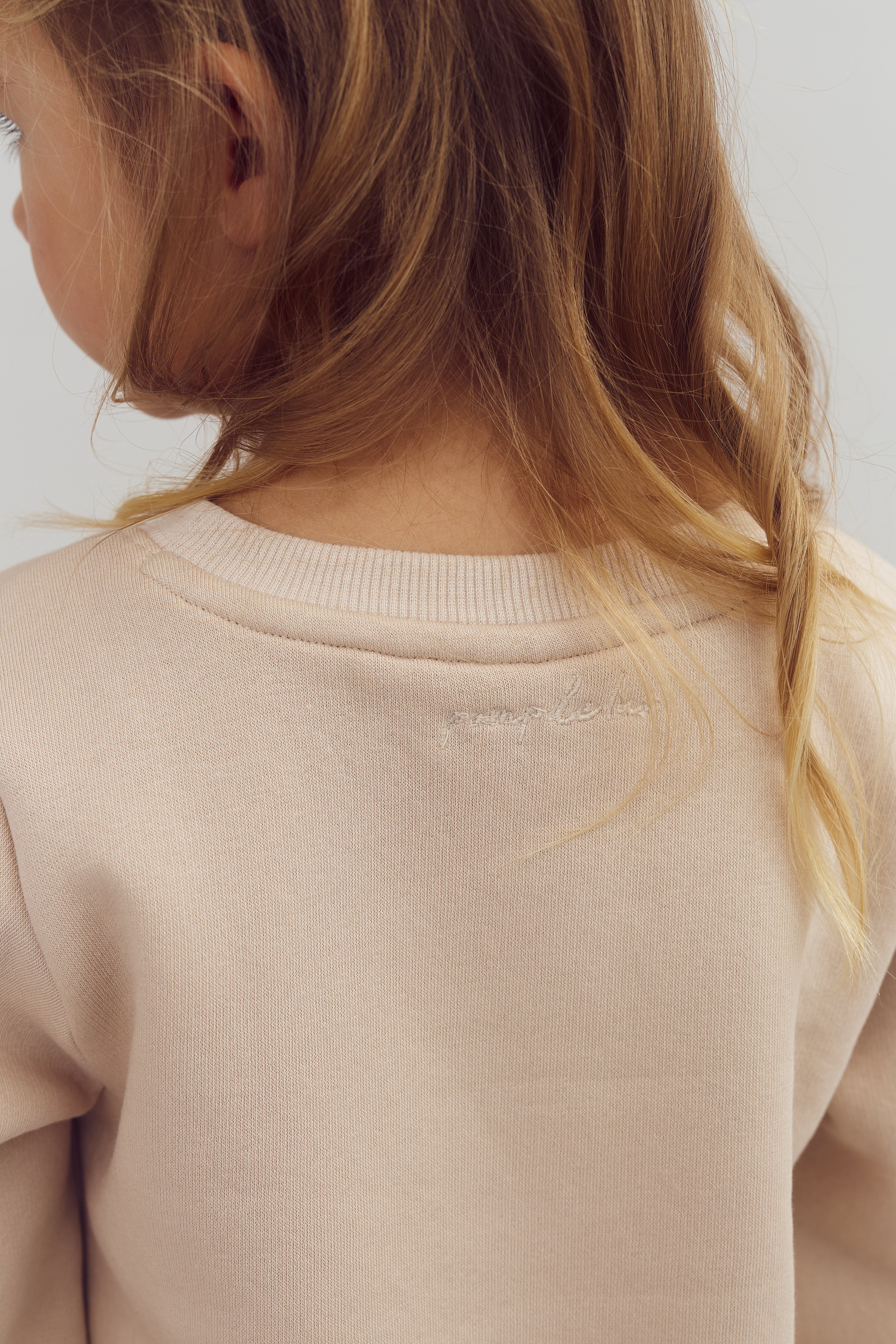 Ein Kind mit hellbraunen Haaren trägt einen beigefarbenen Pullover. Es steht in einer neutralen Umgebung, wobei der Fokus auf der Rückenansicht des Pullovers liegt. Im Text steht: "Jenna & Jesse".