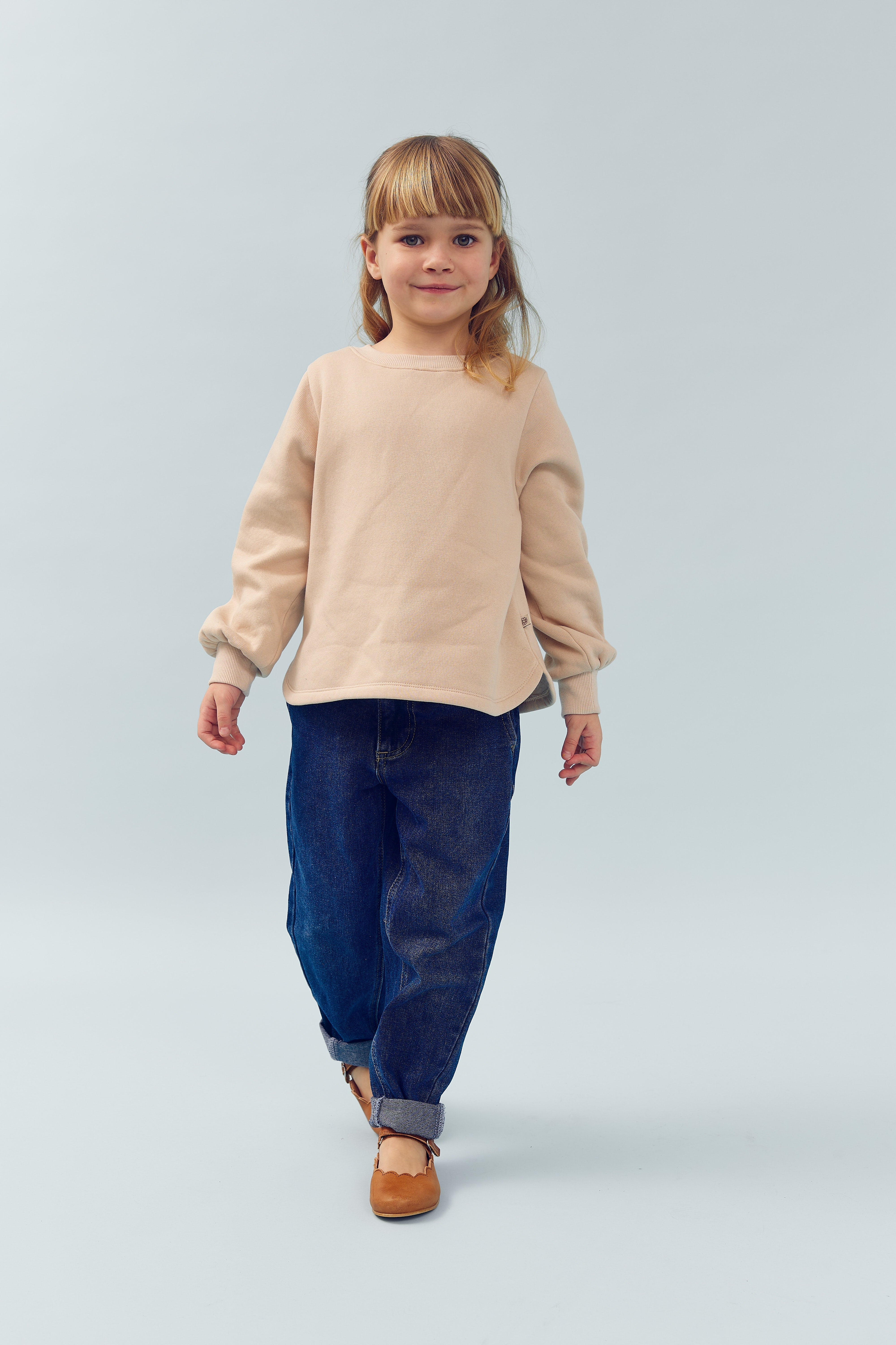Ein Kind trägt einen beigefarbenen Pullover und Jeans, geht aufrecht und selbstbewusst in Richtung Kamera. Der Hintergrund ist einfarbig hellblau.
