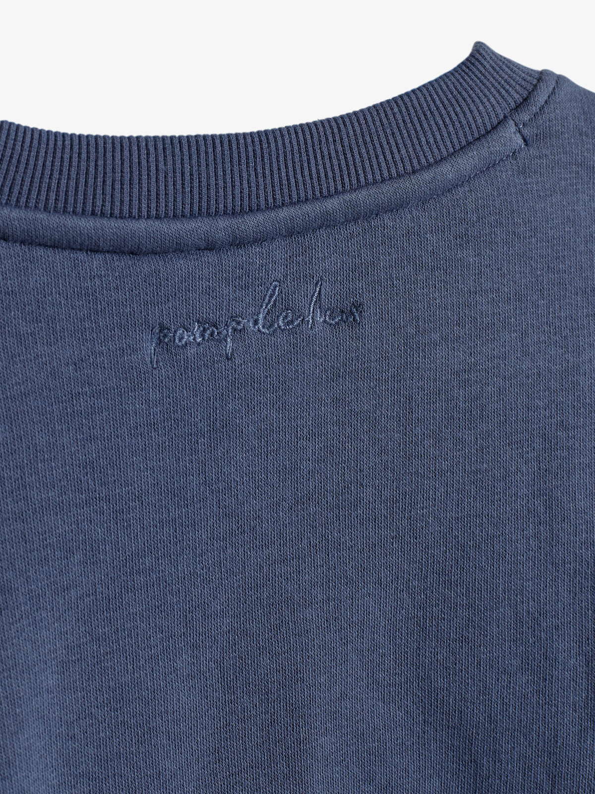 Ein blauer Pullover zeigt am Kragen den Schriftzug "pamplelune". Der Stoff ist gerippt, umgeben von einer gleichmäßigen Textur.