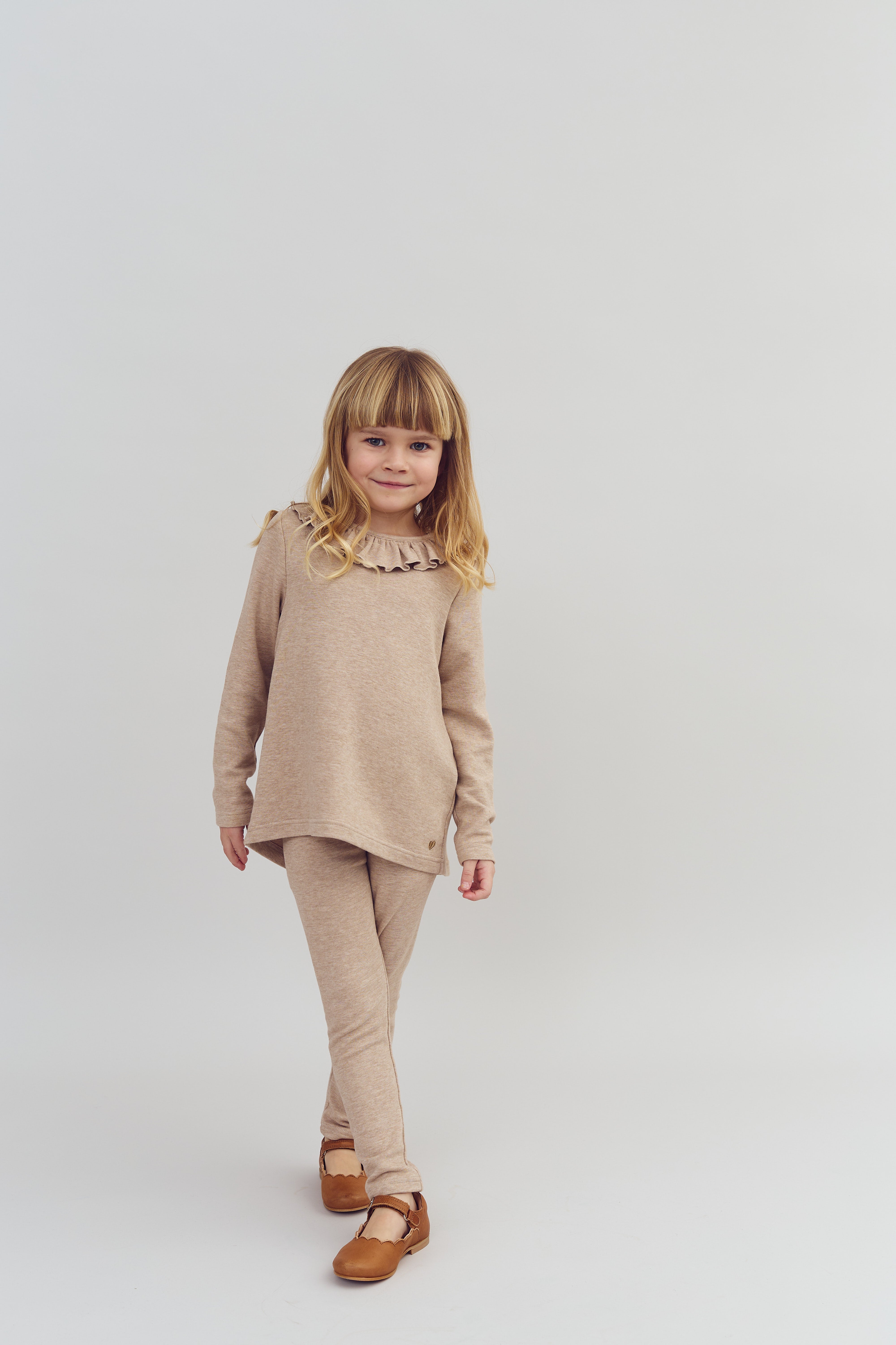 A child in matching beige top and leggings walks confidently, set against a plain, light-colored background. They wear brown shoes and have long, light hair with bangs.