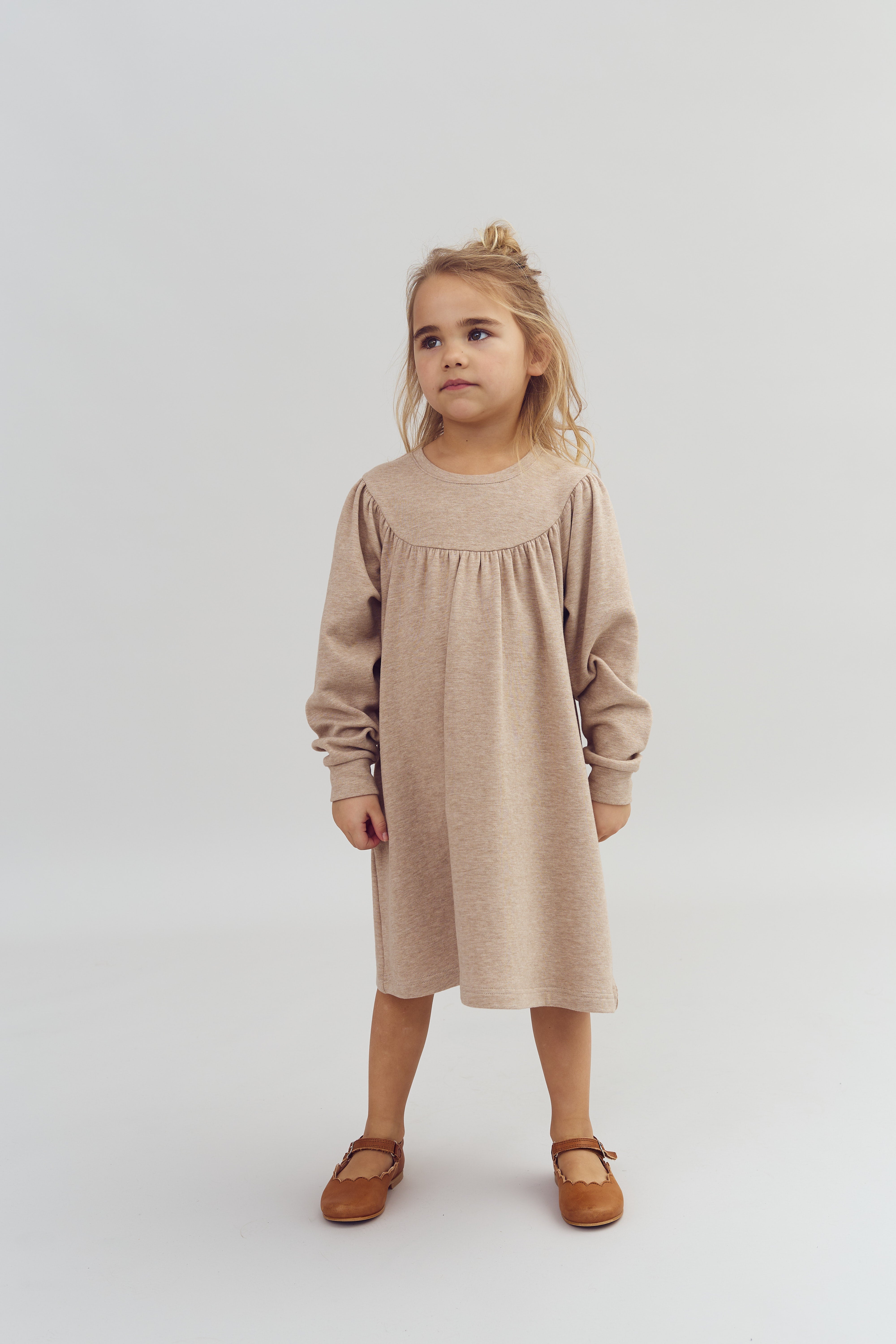Ein Kind steht ruhig, trägt ein beige Kleid und braune Schuhe. Der Hintergrund ist einfarbig hellgrau.
