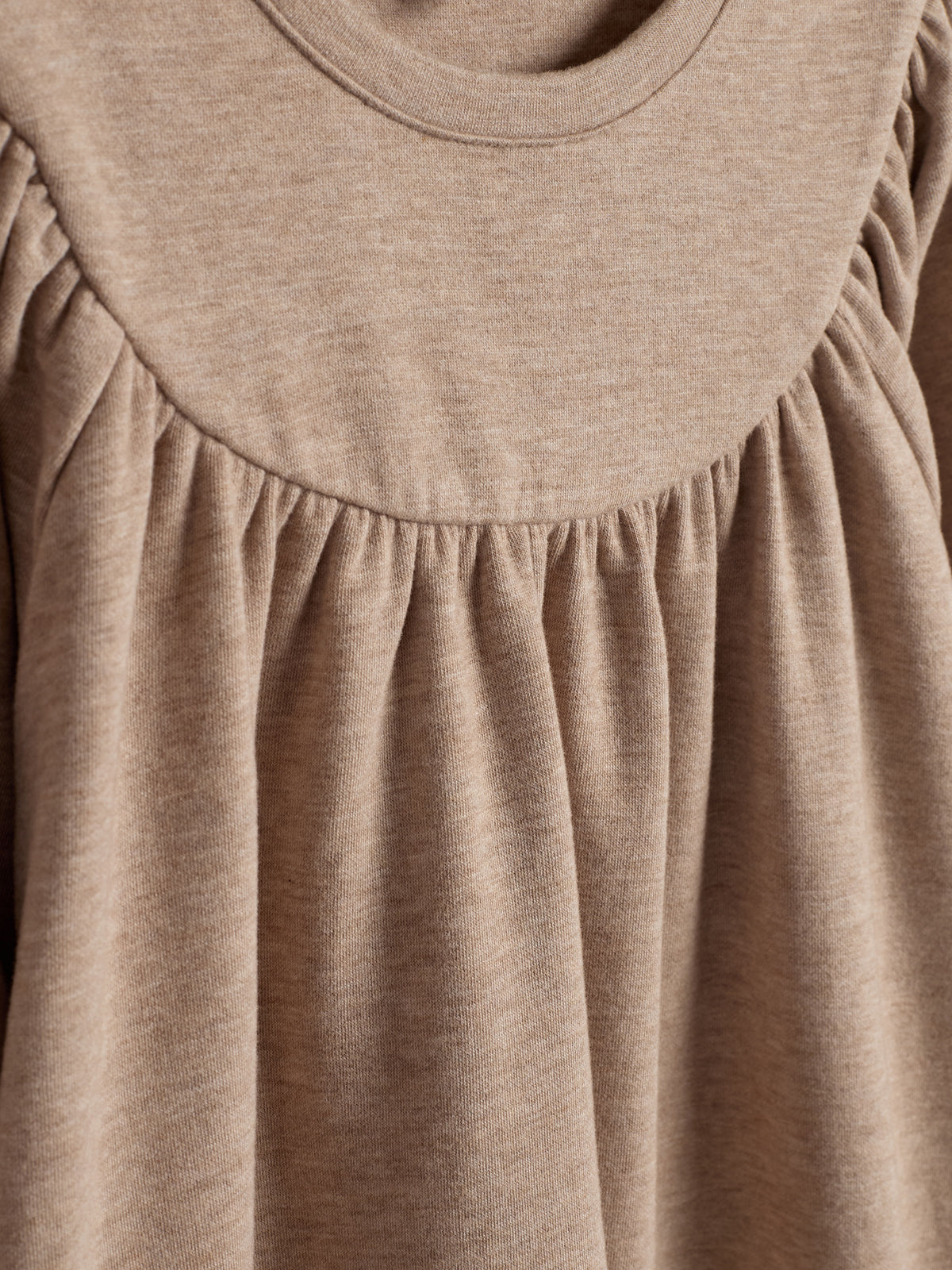 Ein beige Stoff mit gerafften Details und einem runden Halsausschnitt, fließend nach unten, liegt in einem neutralen Hintergrund.