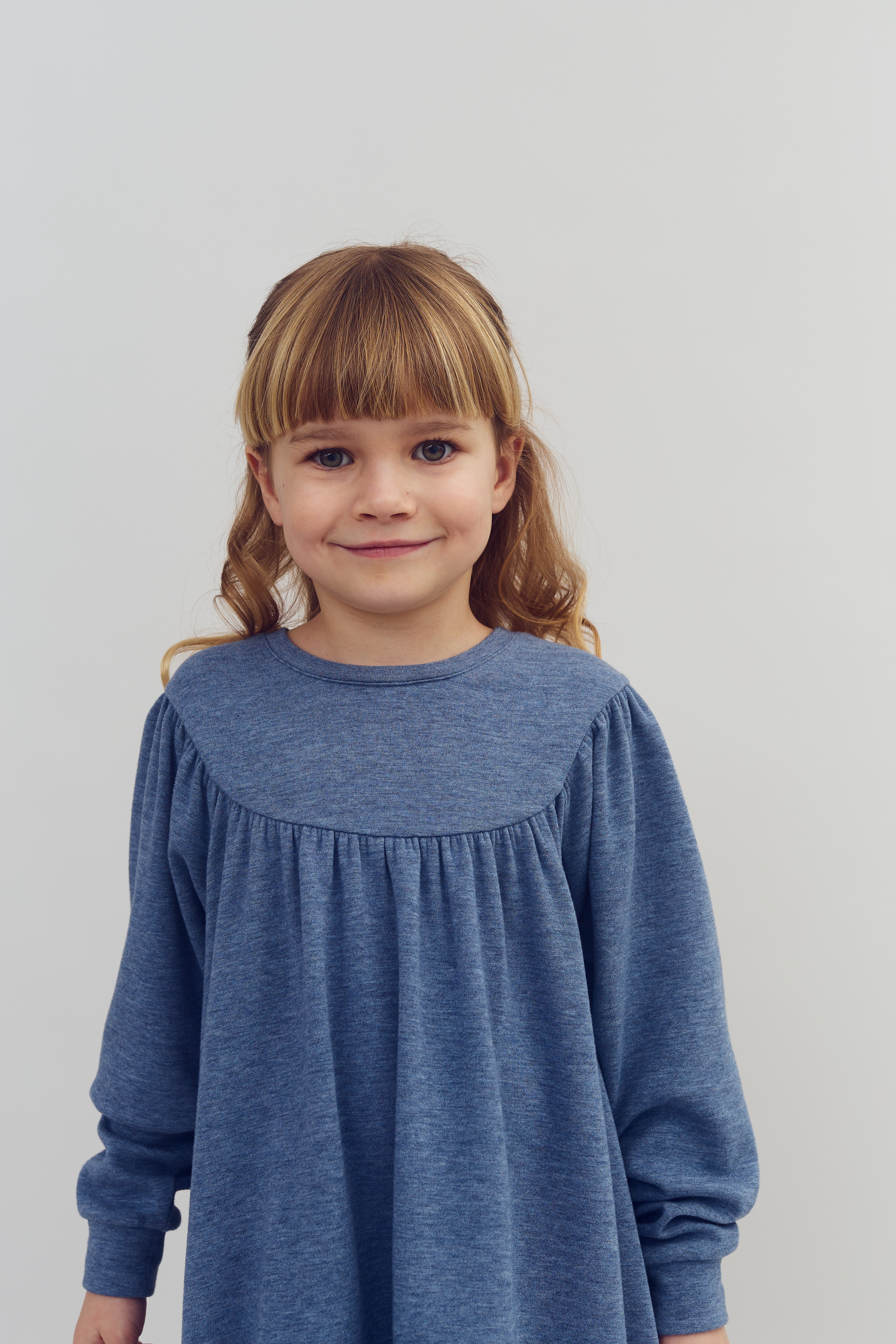 Ein kleines Mädchen steht ruhig und lächelt sanft. Es trägt ein blaues Kleid, während sie vor einem schlichten, hellen Hintergrund steht.