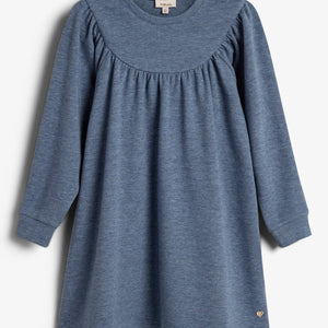Ein blaues langärmeliges Kleid hängt vor einem weißen Hintergrund. Es hat einen runden Ausschnitt und einen gerafften Schulterbereich. Ein kleines goldenes Herz ist am Saum befestigt.