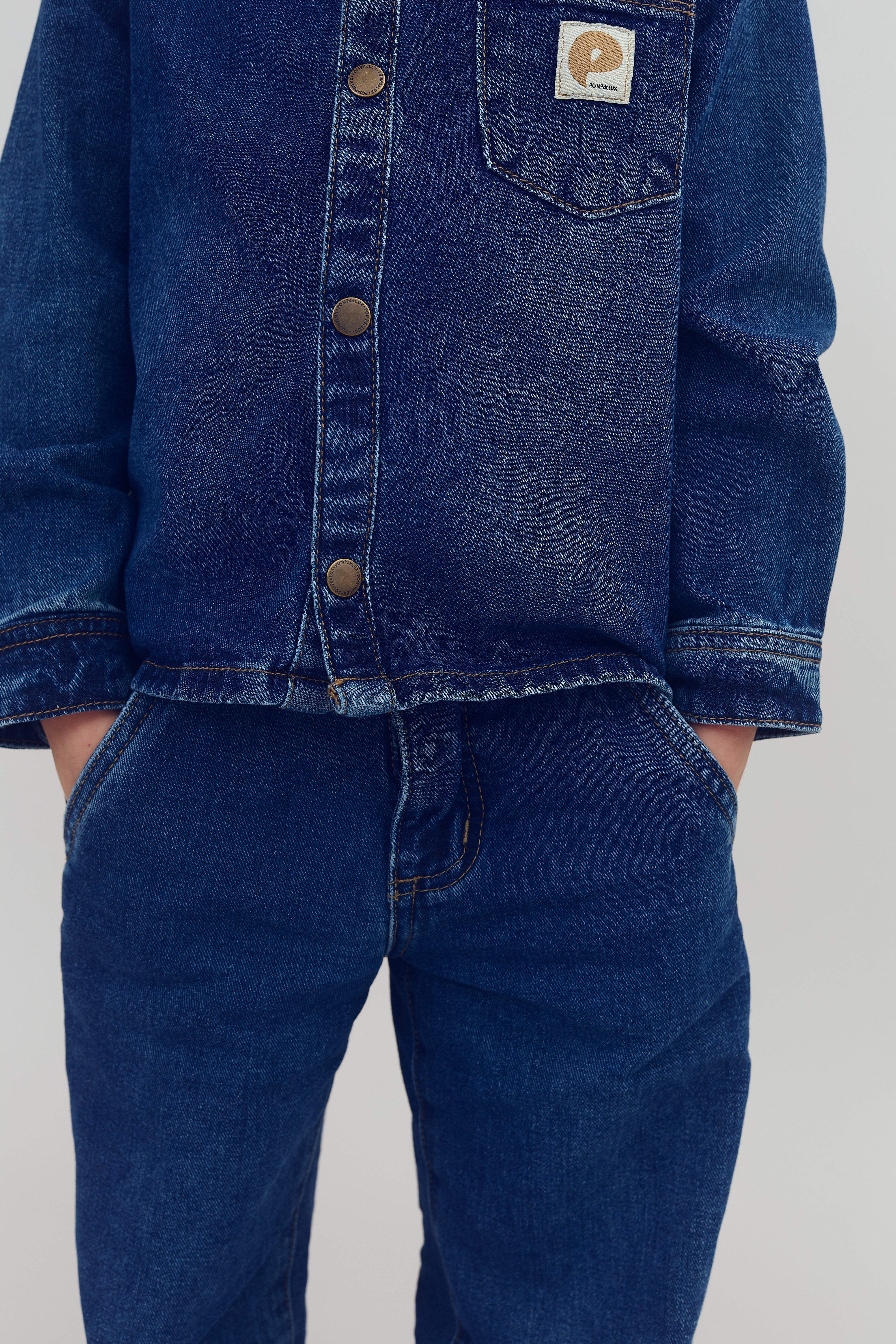 Eine Person trägt eine blaue Jeansjacke und -hose mit den Händen in den Hosentaschen. Auf der Jacke ist ein Etikett mit der Aufschrift "Purewalk". Der Hintergrund ist neutral.