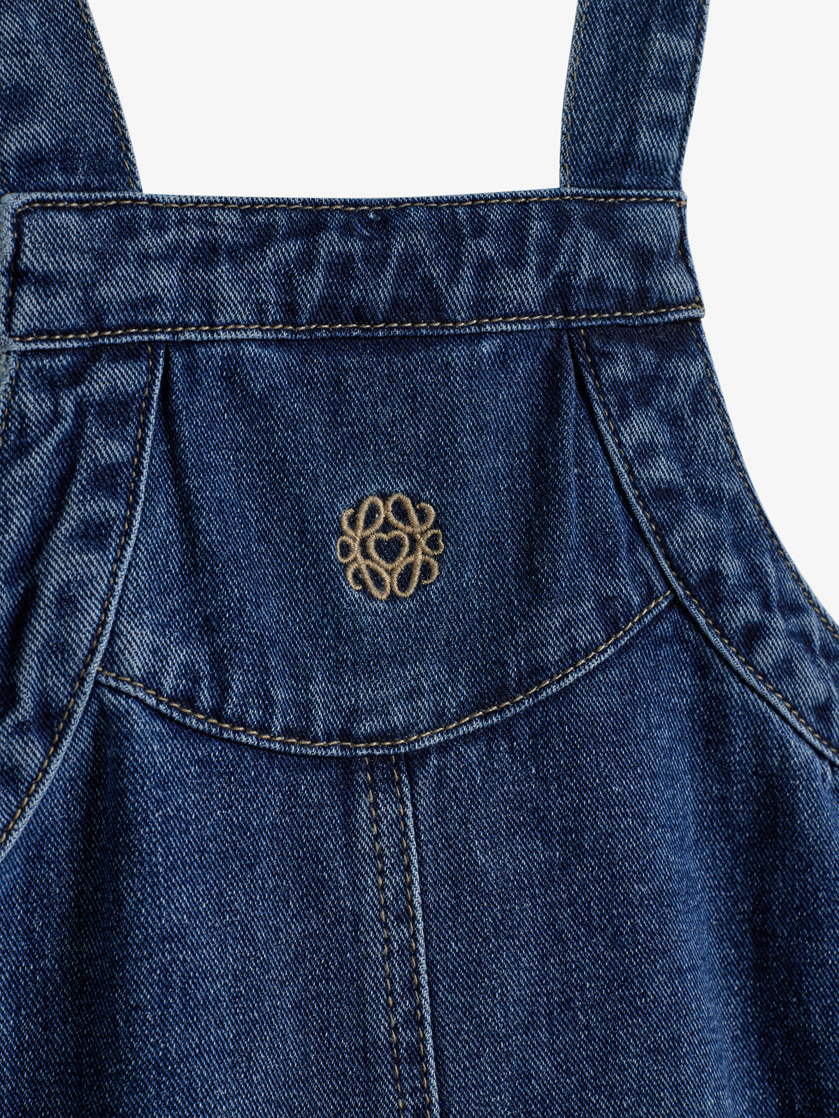 Denimstoff ist mit einem gestickten, dekorativen Muster verziert. Der Kontext zeigt eine Naht und einen ärmellosen Bereich, typisch für Latzhosen oder Schürzen.