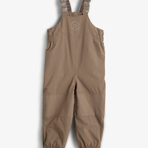 Eine beige Latzhose für Kinder hängt ruhig. Der Stoff hat elastische Beine mit Fußschlaufen. Auf der Latzhose steht: "OUTER ADVENTURE OMPDELUX". Sie hängt vor einem neutralen weißen Hintergrund.