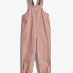 Ein rosa Kinderlatzhose mit verstellbaren, bedruckten Trägern, möglicherweise aus einem wasserabweisenden Material. Die Hose hat einen elastischen Bund an den Beinen und Fußschlaufen. Text: "ULTRAFUNCIONAL AVVENTURA".