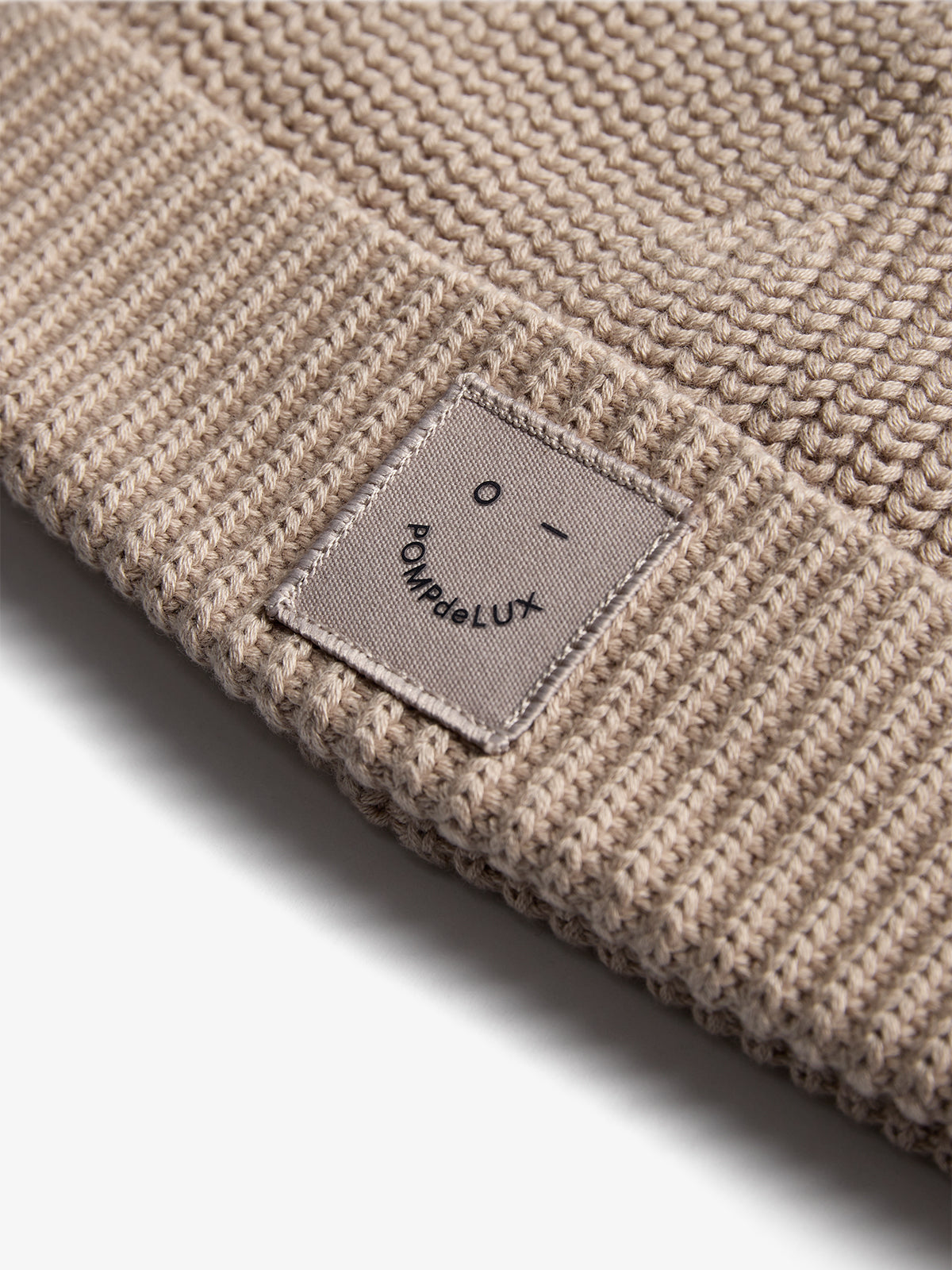A beige knitted fabric with a sewn rectangular patch, displaying a simple face and the text "POMPdeLUX," lies flat. The texture appears detailed and clearly defined.