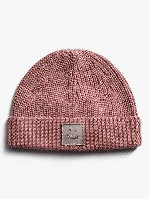 A pink knitted beanie with a folded brim features a stitched patch displaying an abstract face and the text "POMPdeLUX." The beanie rests on a plain white background.