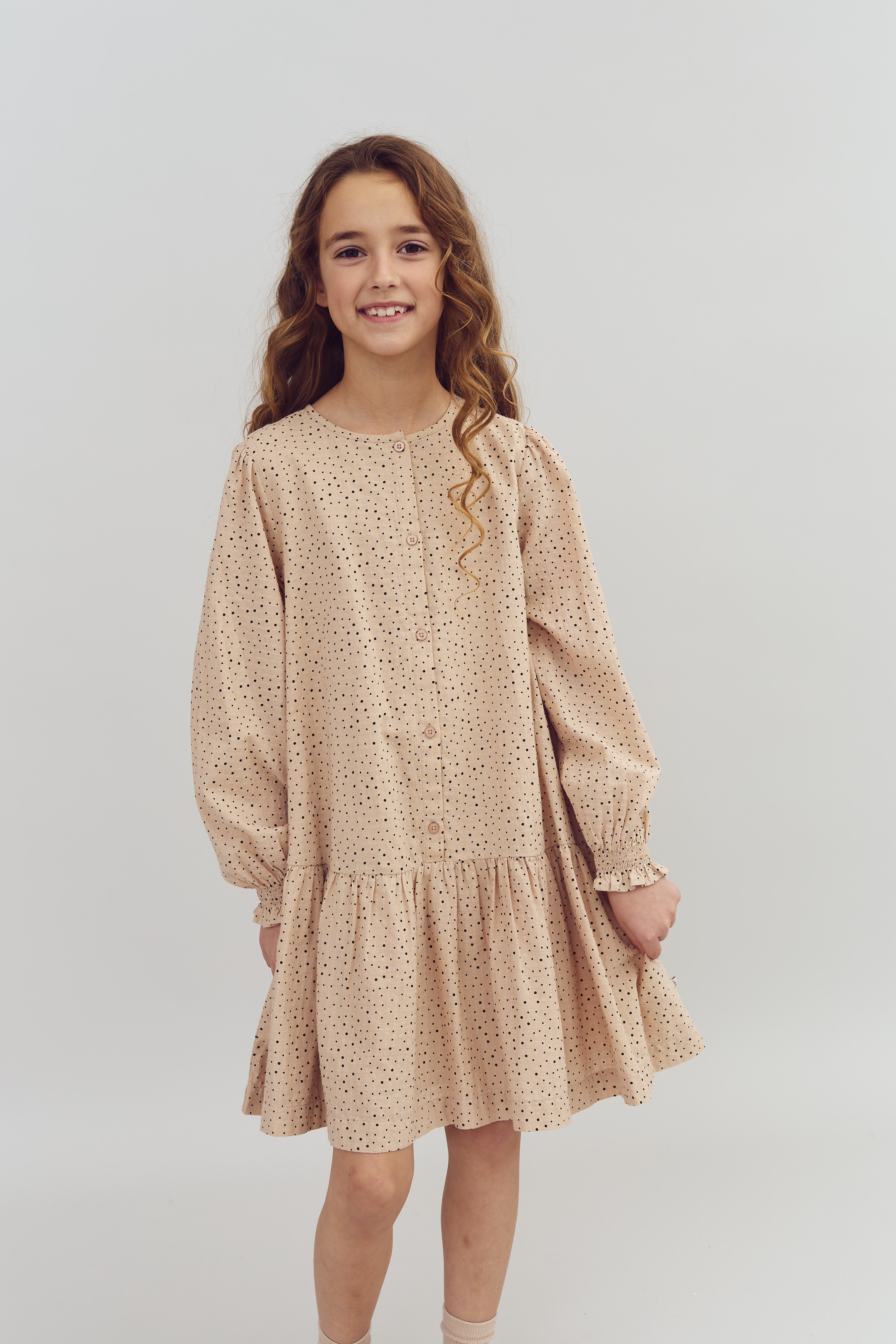 A young girl stands smiling, wearing a beige polka-dotted dress, in a neutral studio setting.