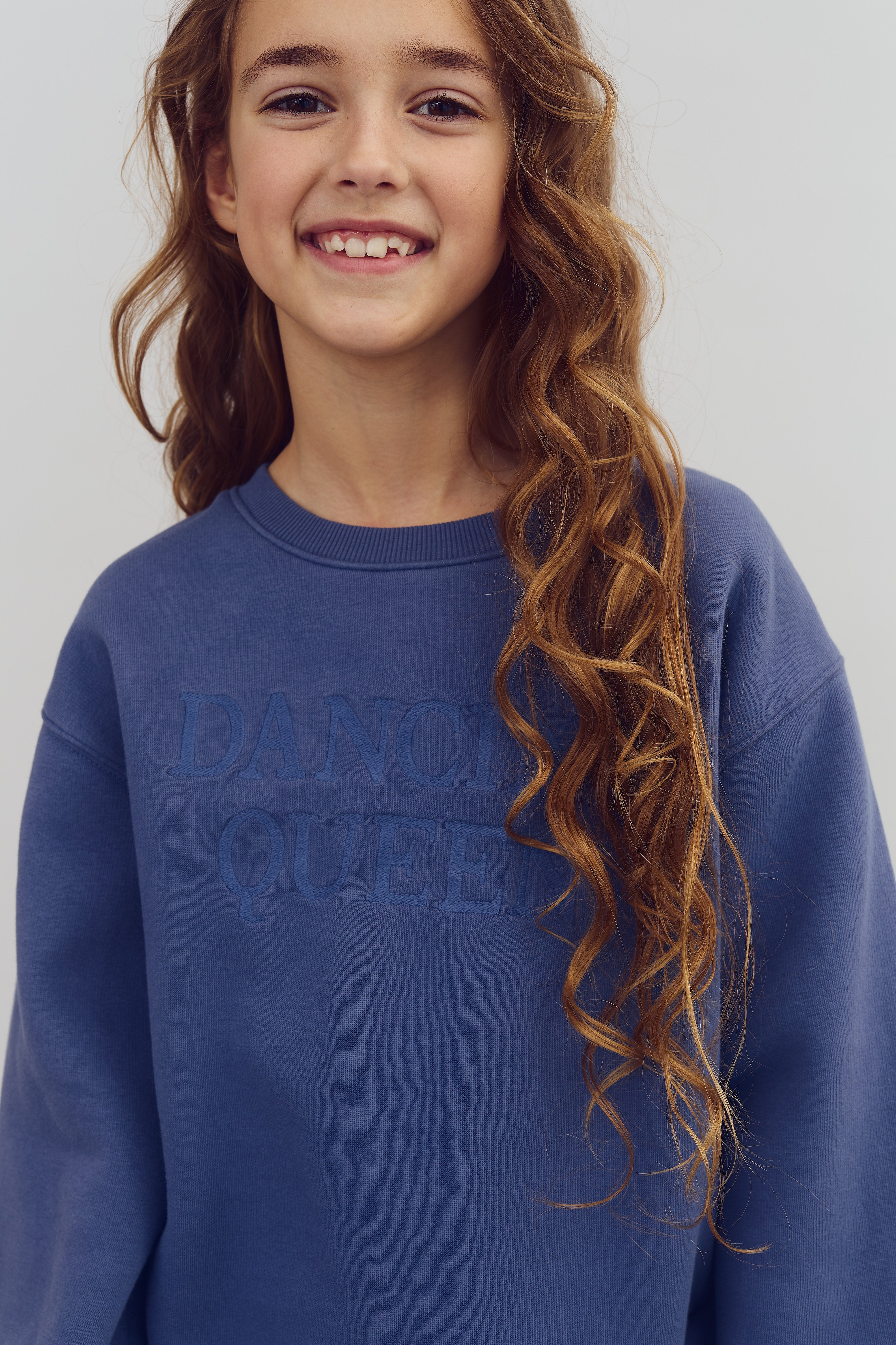 Mädchen lächelt in einem blauen Pullover mit der Aufschrift "DANCING QUEEN" in einer neutralen Umgebung.