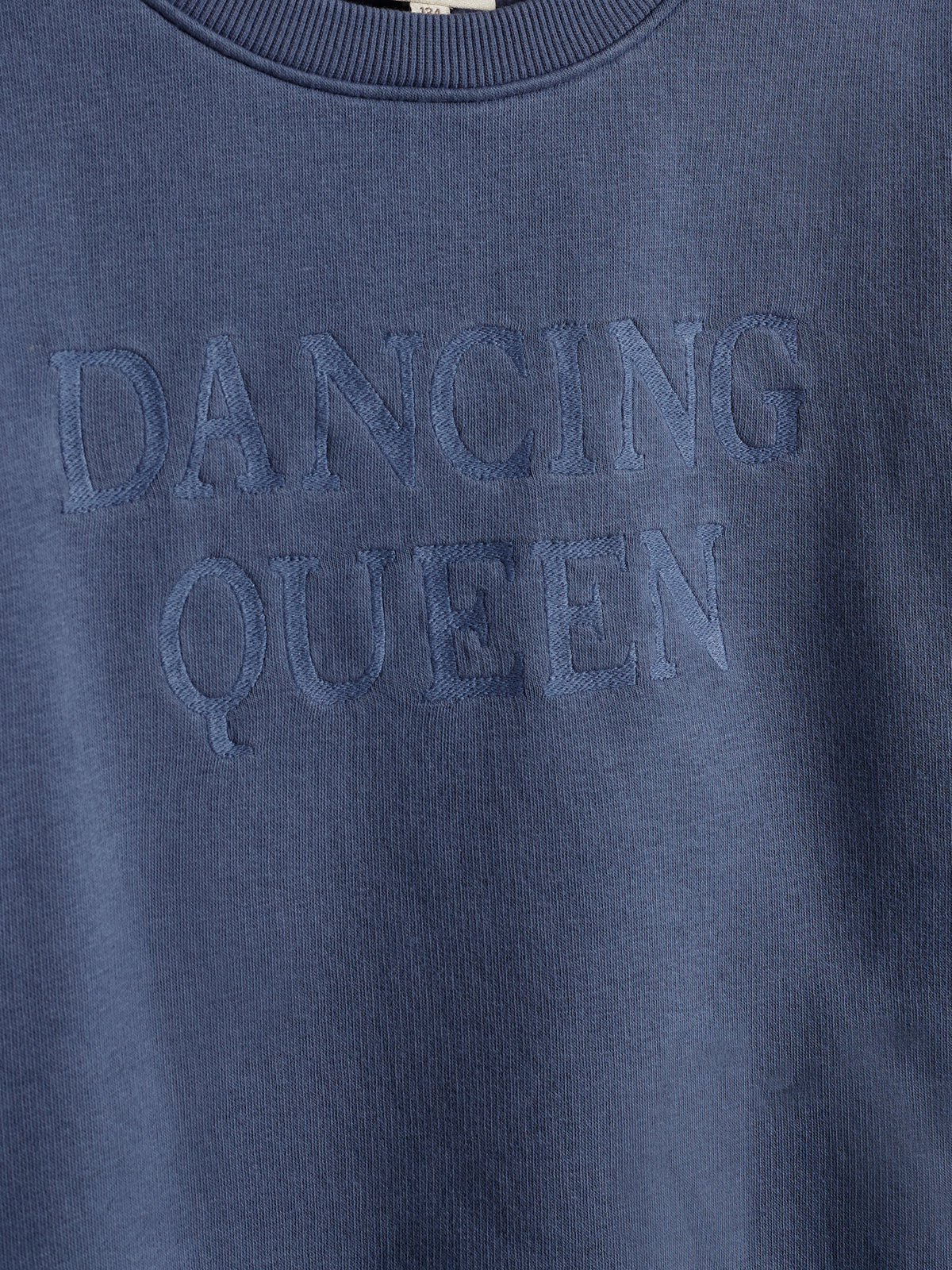 Ein blauer Pullover mit aufgesticktem Text "DANCING QUEEN". Der Stoff wirkt weich und der Pullover hat einen gerippten Rundhalsausschnitt.