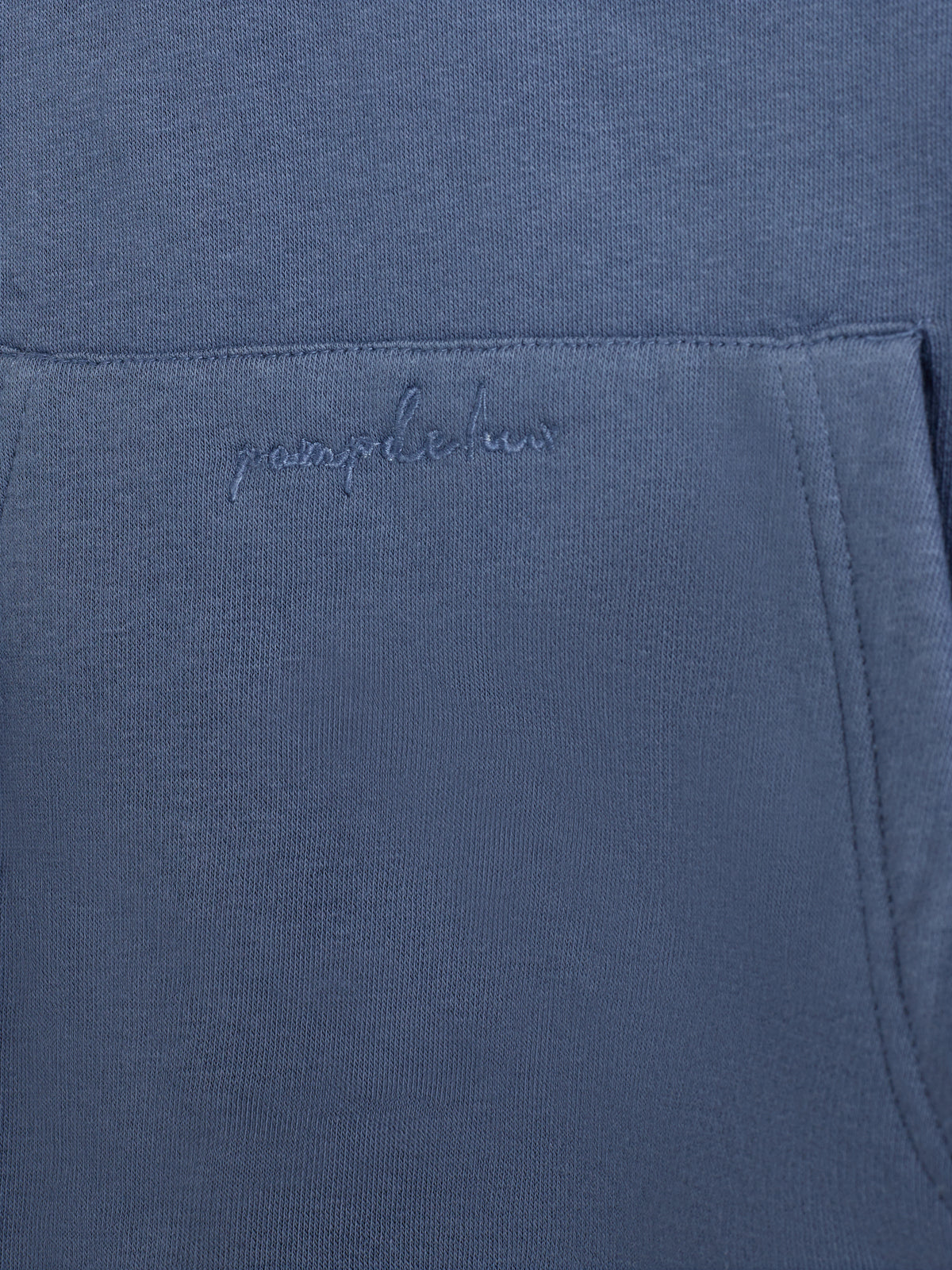 Tasche eines blauen Kleidungsstücks, mit dem Schriftzug "pamplabour" bestickt. Sie befindet sich in der Mitte unter einer Naht, umgeben von gleichmäßiger blauer Stoffstruktur.
