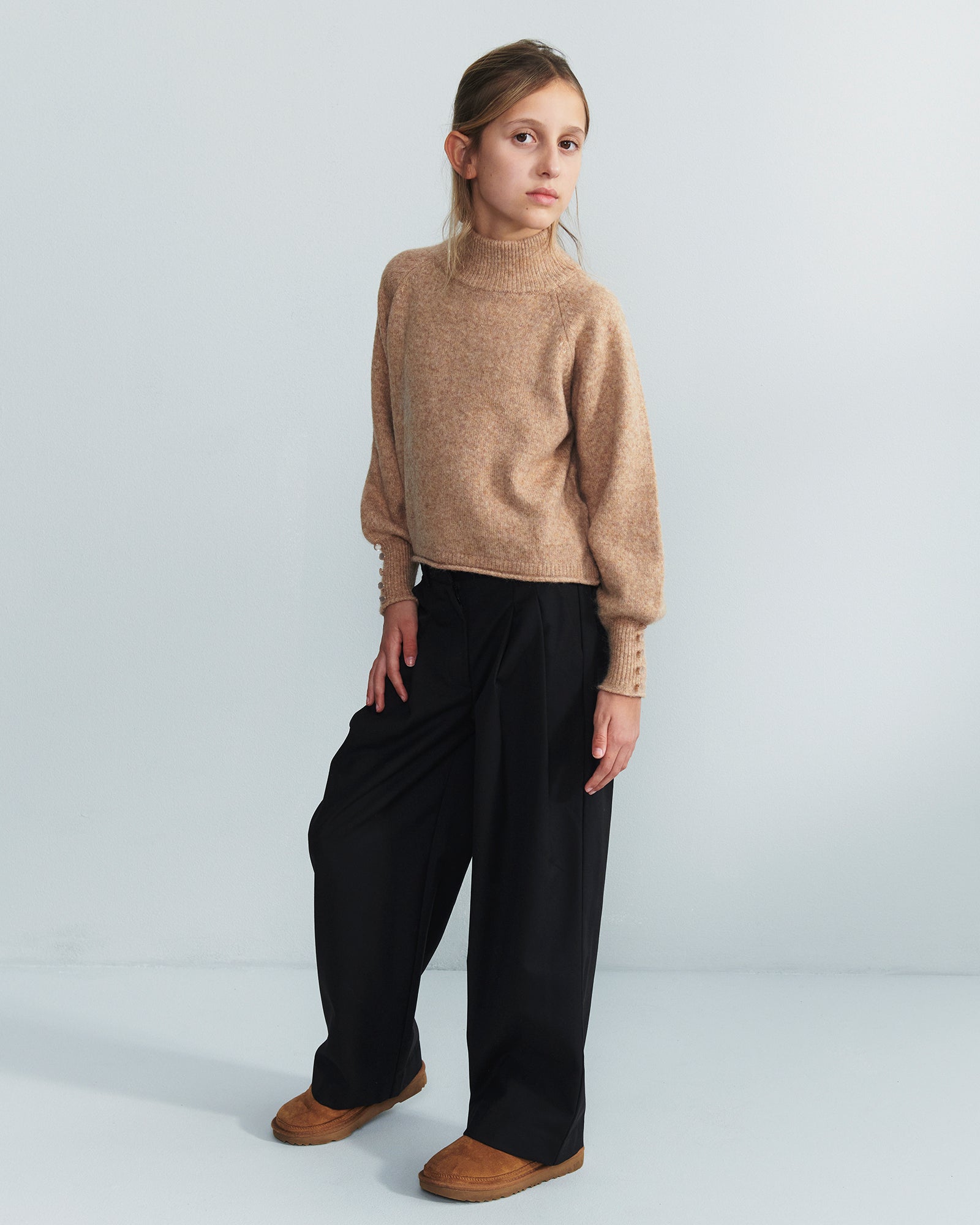 Child standing; wearing a tan turtleneck, wide black pants, and brown shoes; looking at the camera against a plain light gray background.