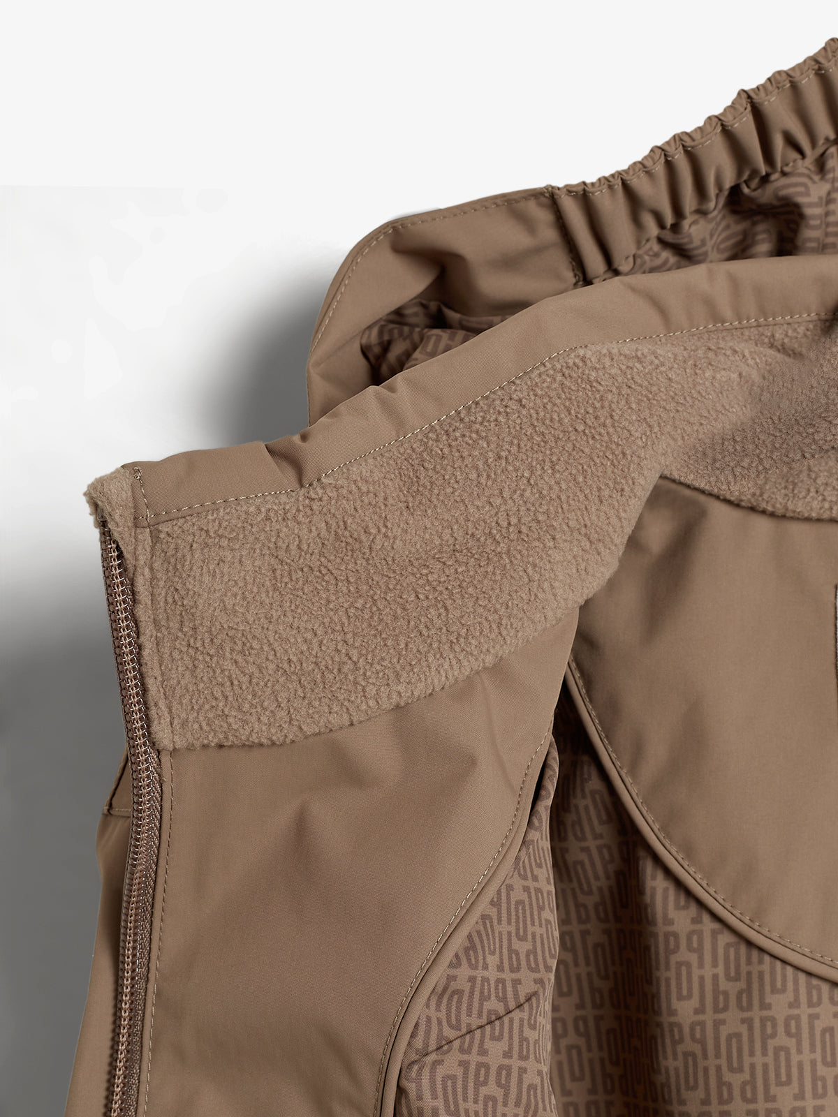 Beigefarbene Jacke mit Fleecekragen, Reißverschluss und einem elastischen Bündchen, liegt auf einer grauen Oberfläche. Das Innenfutter zeigt ein wiederholtes Muster mit den Buchstaben „P“ und „B“.
