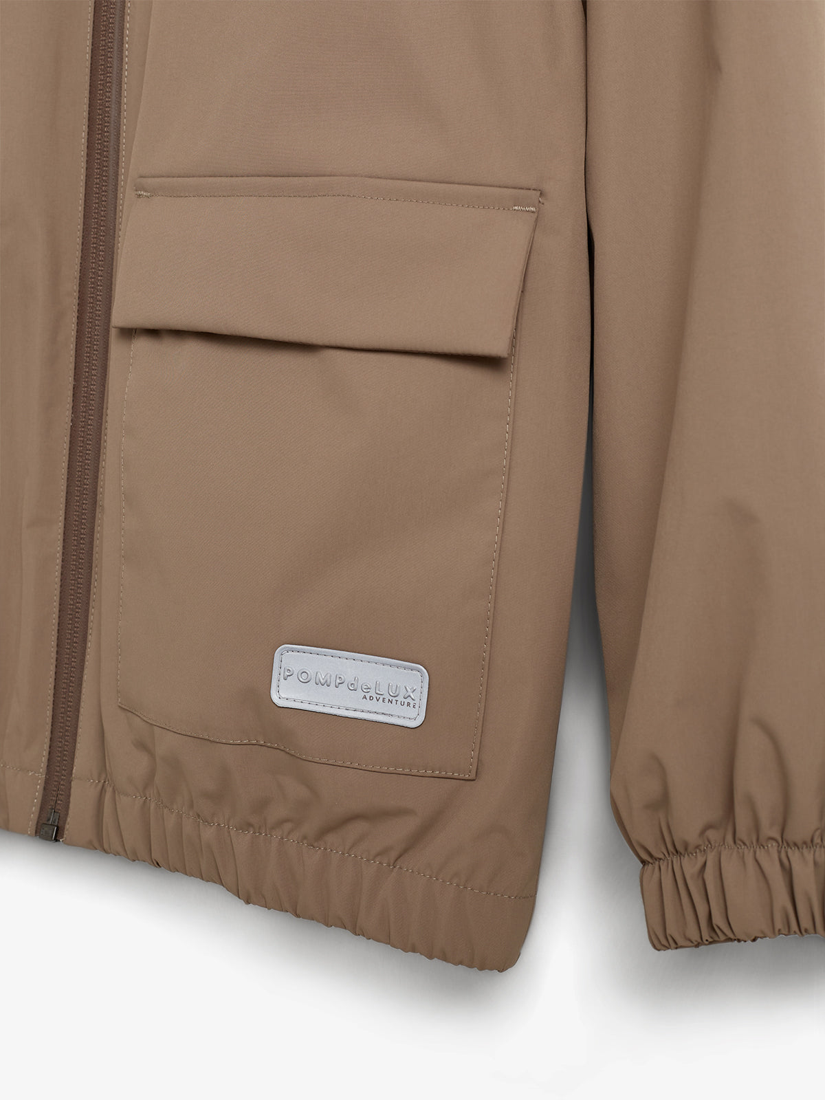 Beigefarbene Jacke mit Reißverschluss und Tasche. Auf der unteren Tasche befindet sich ein Label mit der Aufschrift „POMPdeLUX ADVENTURE“. Elastische Bündchen am unteren Rand und Ärmeln.