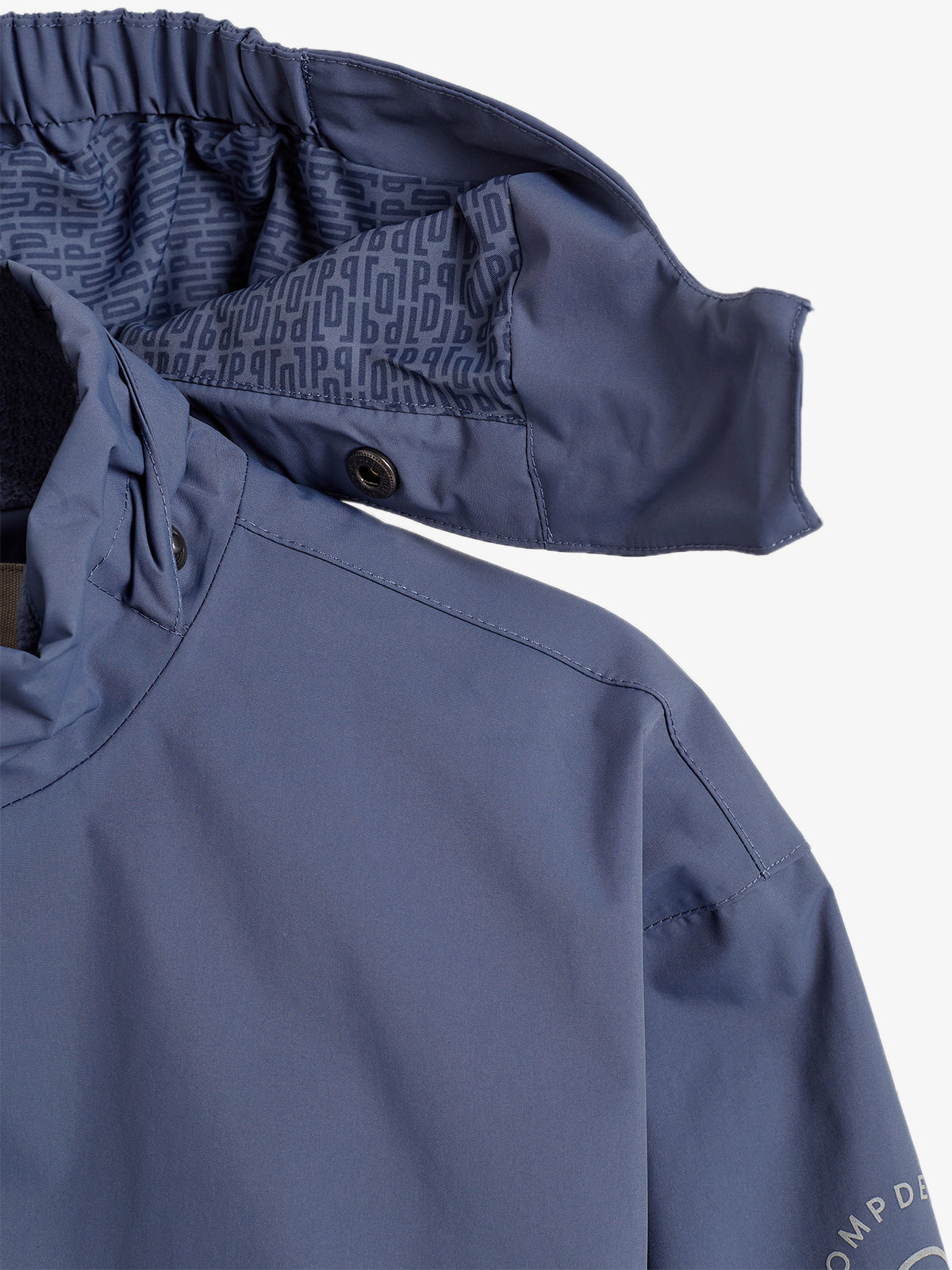 Eine blaue Jacke mit hochstehendem Kragen, zeigt einen unverschlossenen Kragen mit einem elastischen Einsatz. Innenfutter hat ein sich wiederholendes Muster aus stilisierten Buchstaben. Text: "DES COMP".