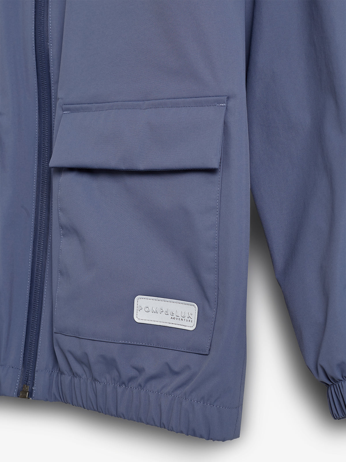 Eine blaue Jacke mit Reißverschluss und großer Fronttasche ist zu sehen. Ein Etikett auf der Tasche trägt die Aufschrift "POMPdeLUX ADVENTURE". Die Jacke hat elastische Bündchen.