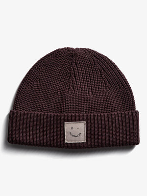 A knitted beanie resting flat on a white surface features a visible patch with a smiley face and "POMPdeLUX" text on the folded brim.